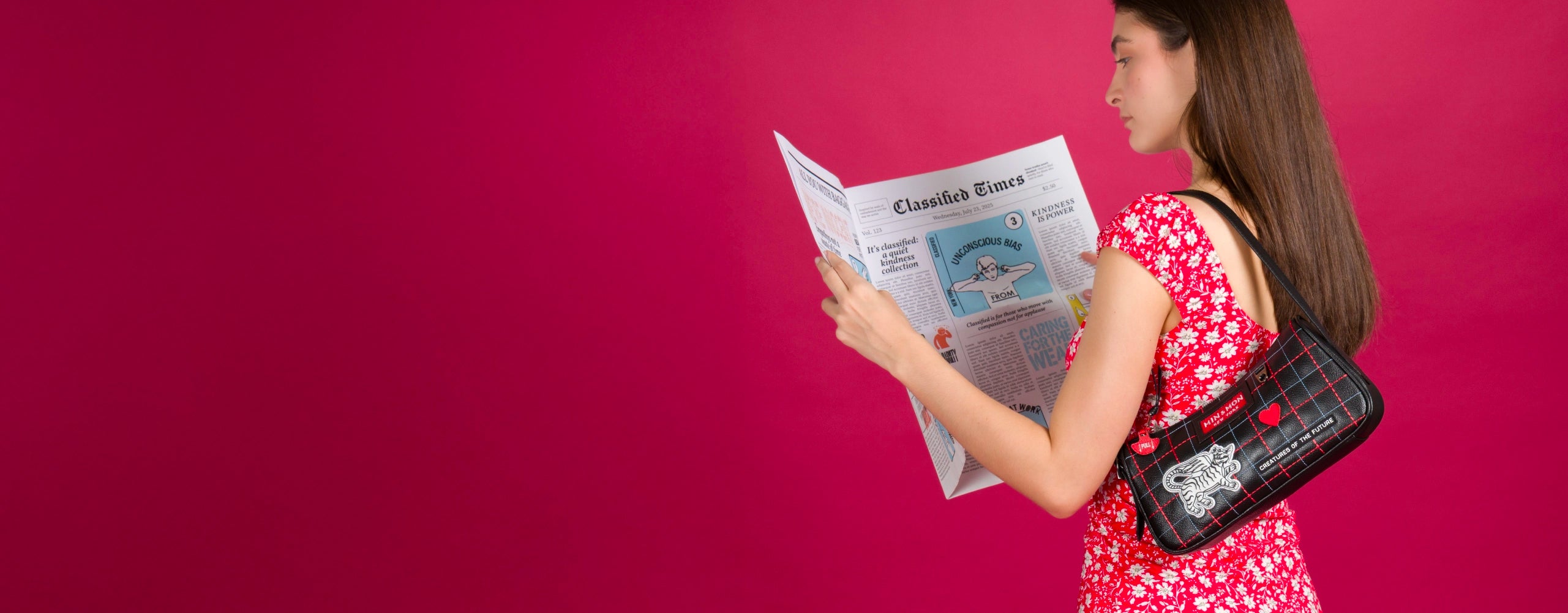 A woman in a red floral dress stands against a bold pink background, reading a whimsical newspaper titled “Classified Times.” She carries a black shoulder bag with red grid lines, embroidered details including a winged tiger, hearts, and the words “Creatures of the Future.”