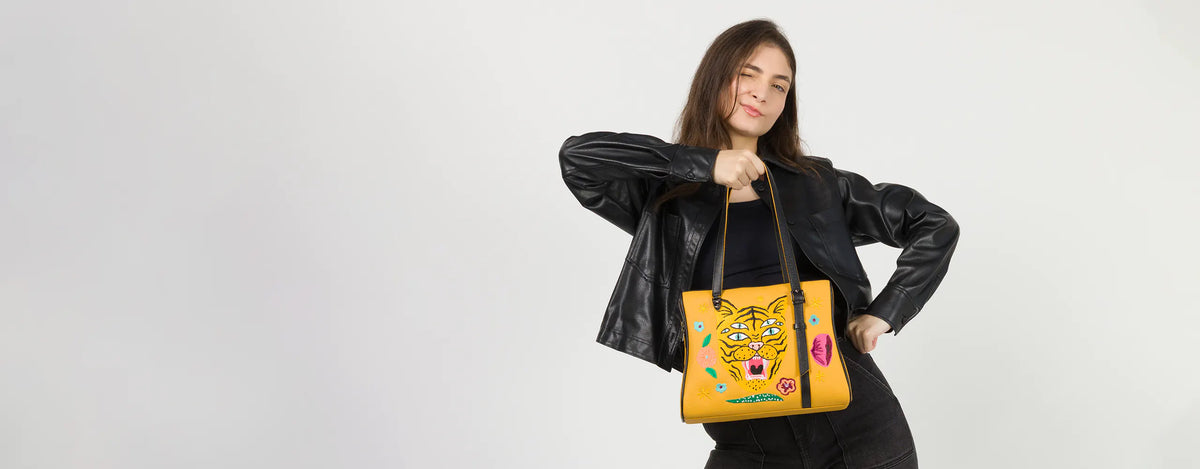 Model winking in a black leather jacket, holding the Min and Mon Esther Happy Tiger handbag in yellow leather featuring a colorful embroidered tiger from the Creatures of the Future collection. Light gray background.