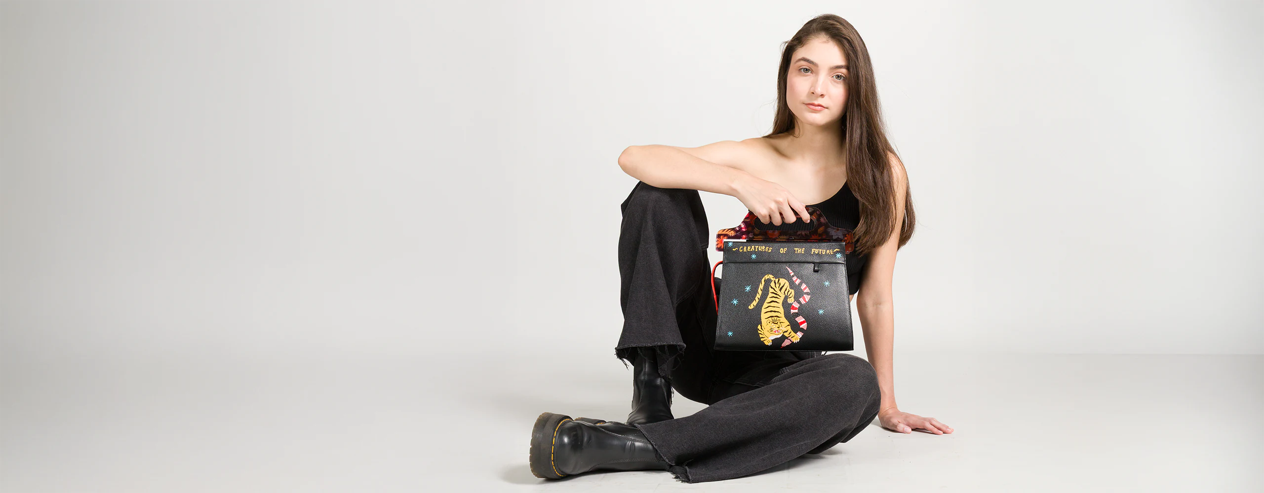 Model seated in studio holding a black Min & Mon handbag with embroidered tiger and “Creatures of the Future” message.