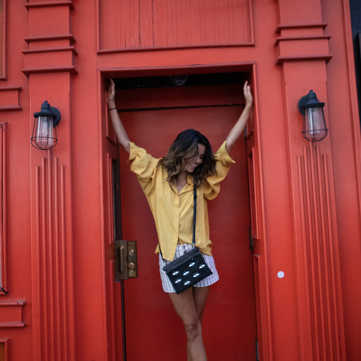Woman posing in front of a bold red doorway wearing a black eye-detail crossbody bag.