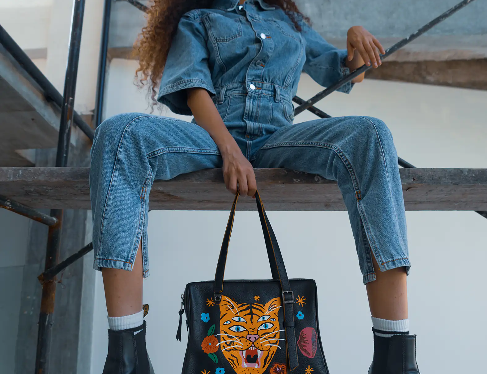 Woman with curly hair sits on a metal stair structure wearing a denim jumpsuit and black boots, holding a black tote bag featuring a colorful embroidered tiger face design.