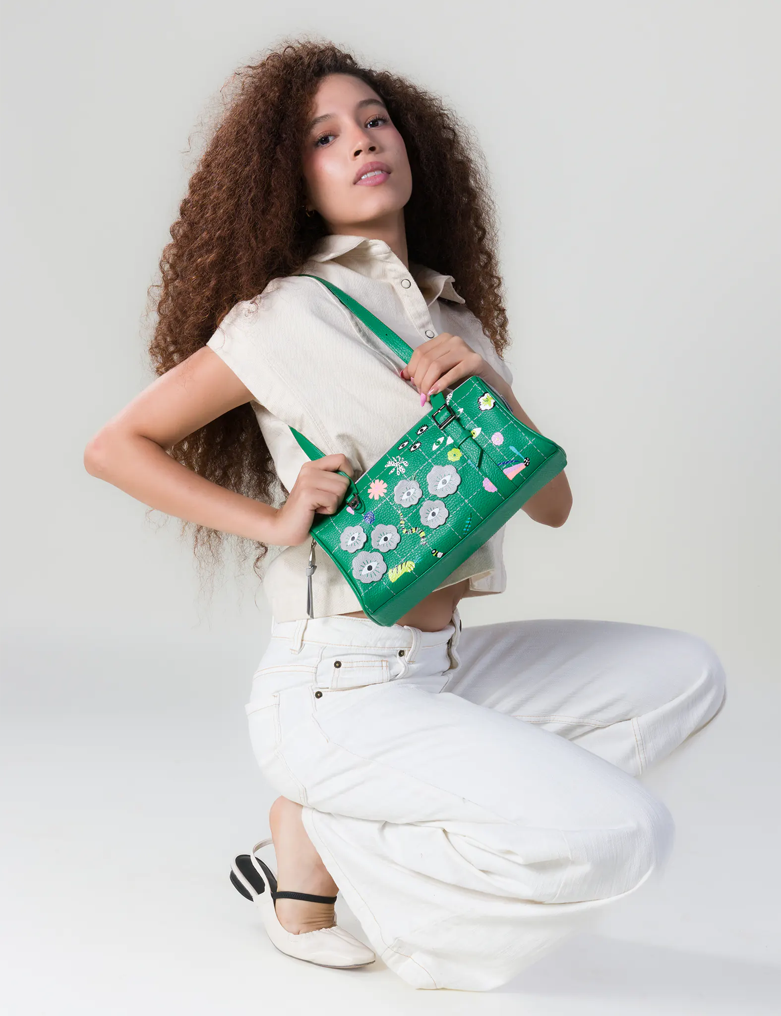 Woman with curly hair kneels in a studio, wearing a cream top and white pants, holding a green handbag decorated with colorful eye motifs.