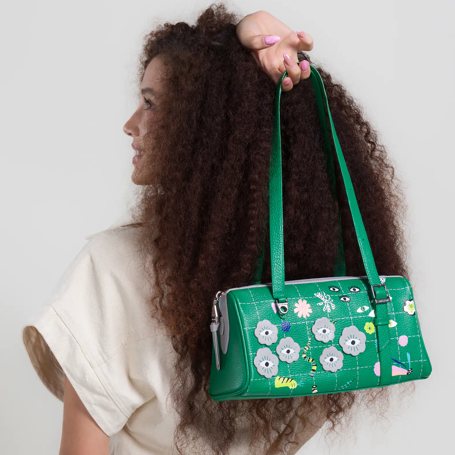 Woman with curly hair turns sideways, holding a green shoulder bag with playful eye designs, wearing a light top against a neutral background.