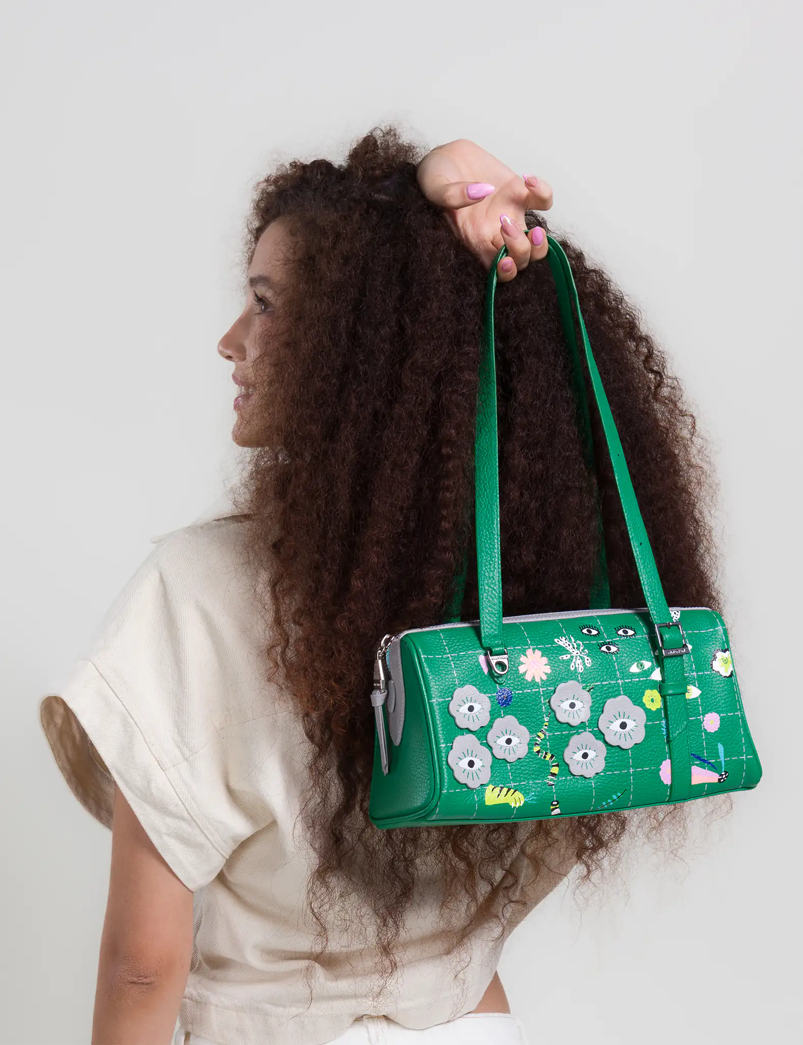 Woman with curly hair turns sideways, holding a green shoulder bag with playful eye designs, wearing a light top against a neutral background.