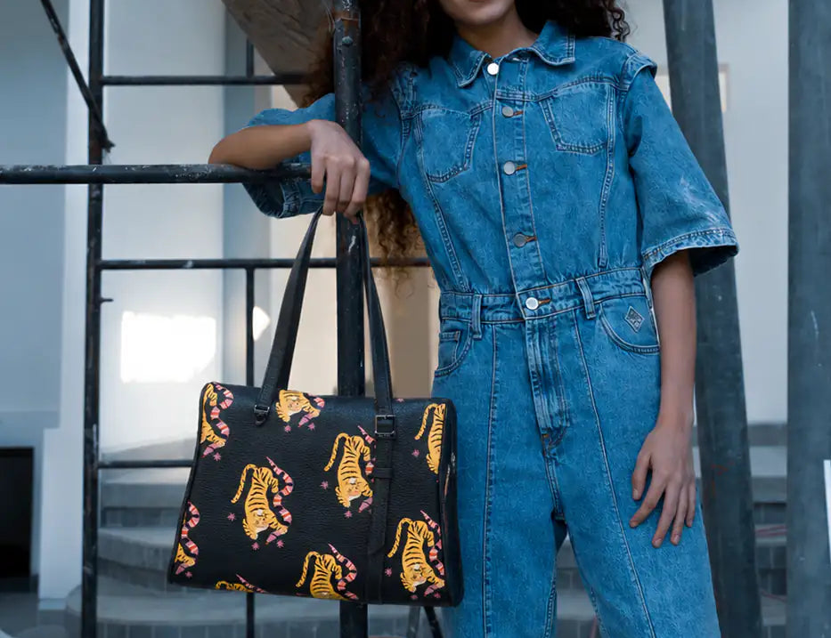 Smiling woman in a blue denim jumpsuit poses by scaffolding in a building under renovation, holding a black tote with yellow tiger prints and pink accents.