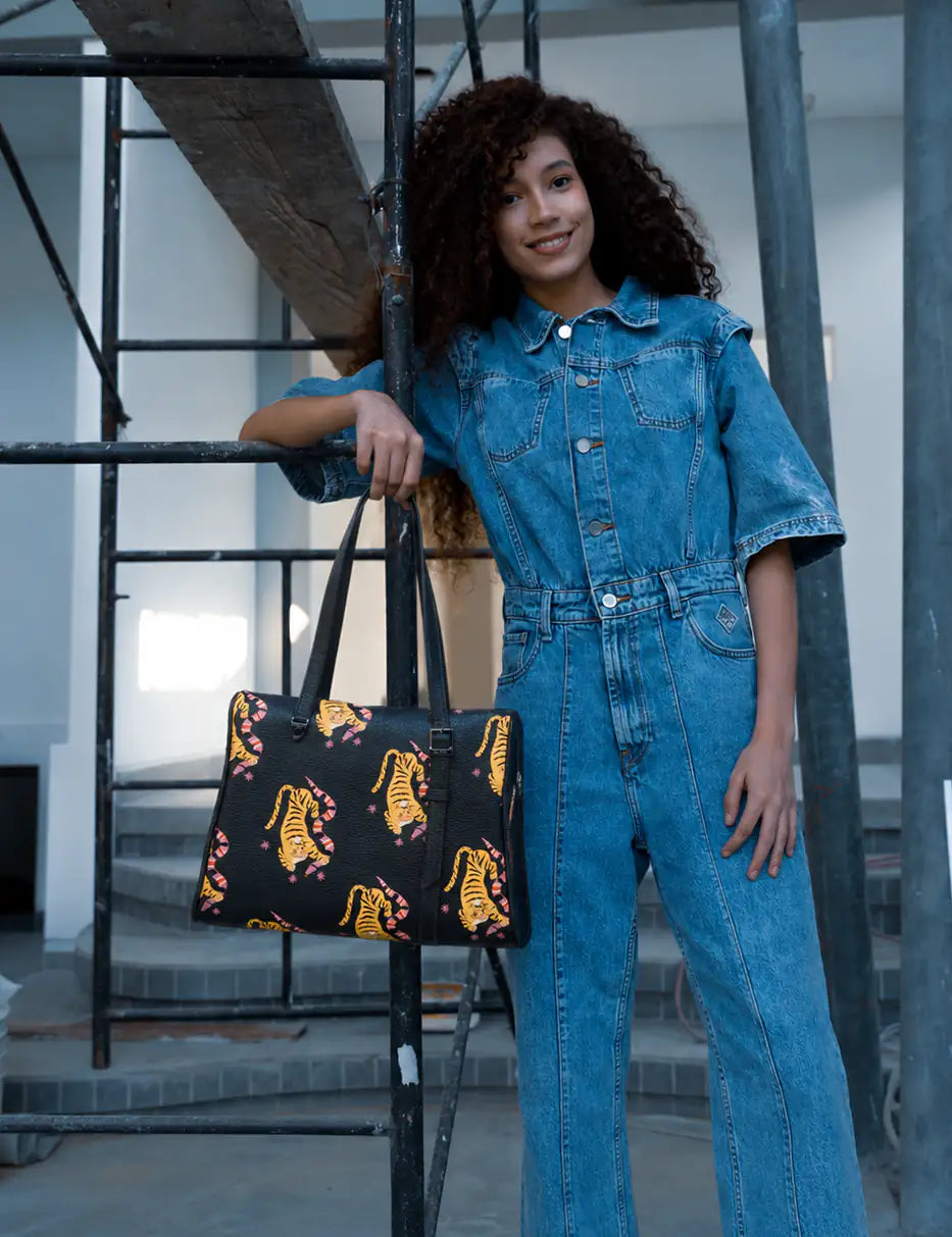 Smiling woman in a blue denim jumpsuit poses by scaffolding in a building under renovation, holding a black tote with yellow tiger prints and pink accents.