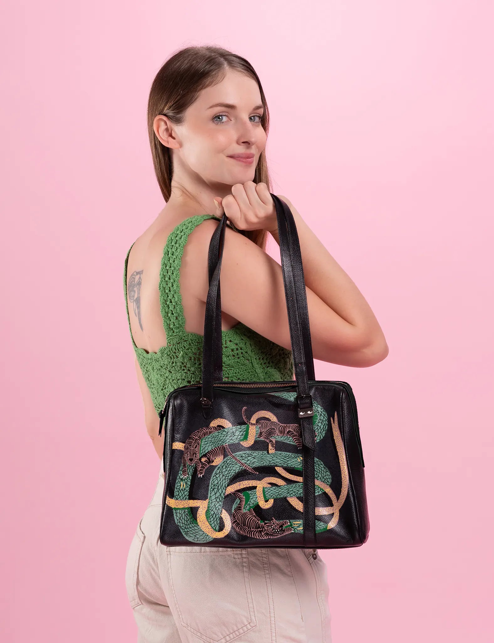 A woman with light skin and long brown hair is posing against a pink background. She is wearing a green sleeveless top with a crochet-like texture and light-colored pants. She has a tattoo on her left shoulder blade and is holding an "Esther Black Duffle Bag - Tangle Tales Print" with two long straps. The handbag features a colorful print design of green snakes intertwined with tigers. The woman is smiling slightly and looking over her right shoulder at the camera.