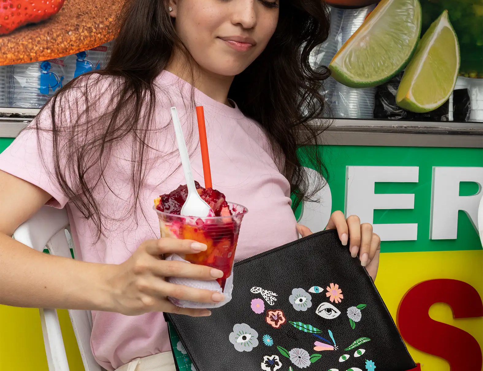 A woman in a pink shirt and white pants smiles slightly while holding a fruity dessert with a spoon and straw. She carries a black clutch with colorful floral patterns, sitting against a vibrant background with fruit imagery, creating a cheerful and relaxed vibe.