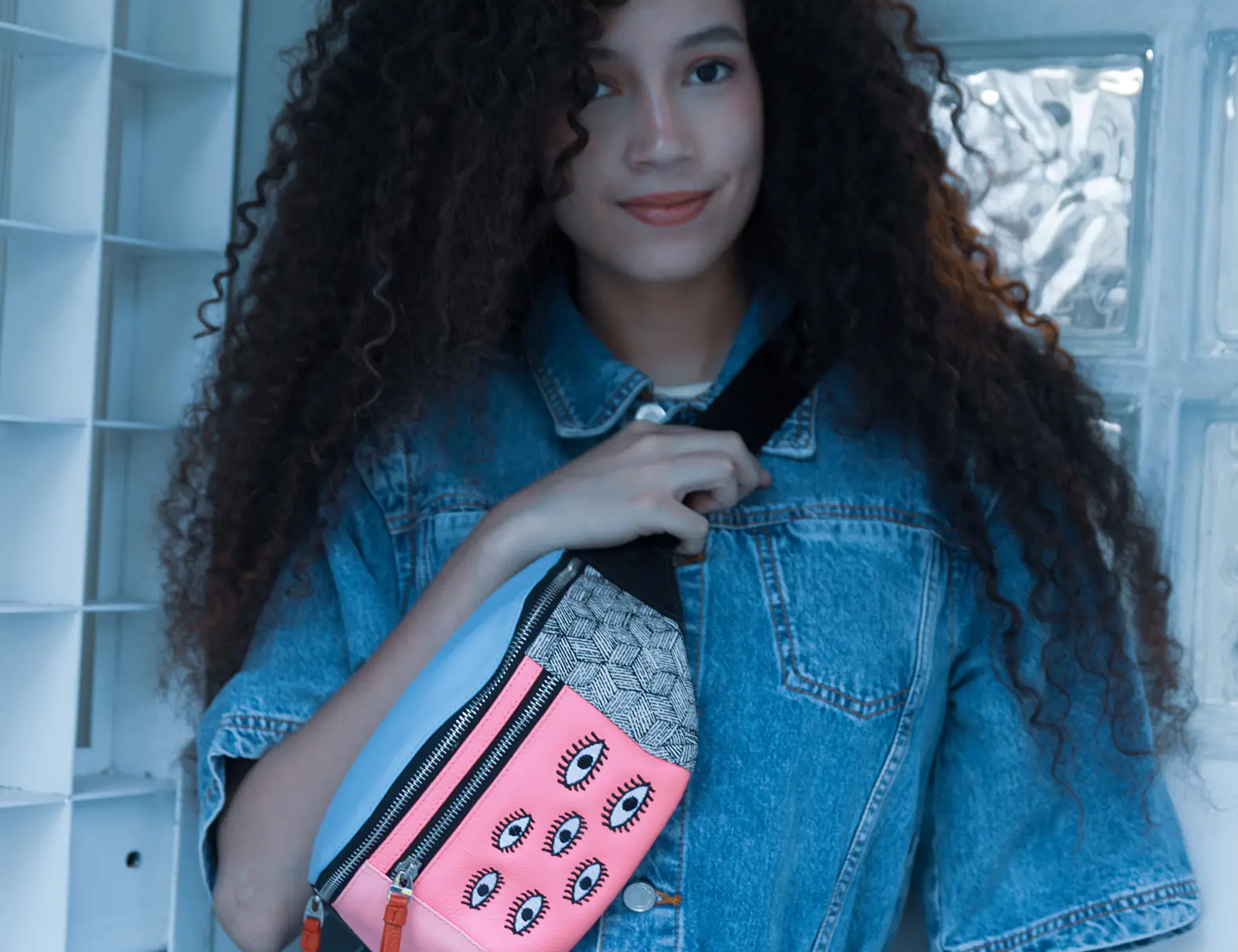 Woman with curly hair in a denim outfit wears a colorful fanny pack with pink panel and eye motifs, posing indoors against a light blue tiled wall.