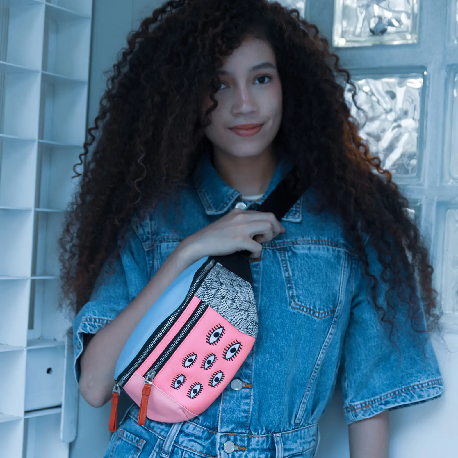Woman with curly hair in a denim outfit wears a colorful fanny pack with pink panel and eye motifs, posing indoors against a light blue tiled wall.
