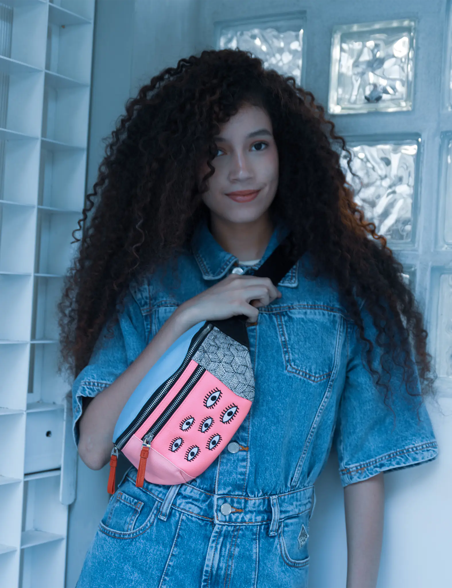 Woman with curly hair in a denim outfit wears a colorful fanny pack with pink panel and eye motifs, posing indoors against a light blue tiled wall.