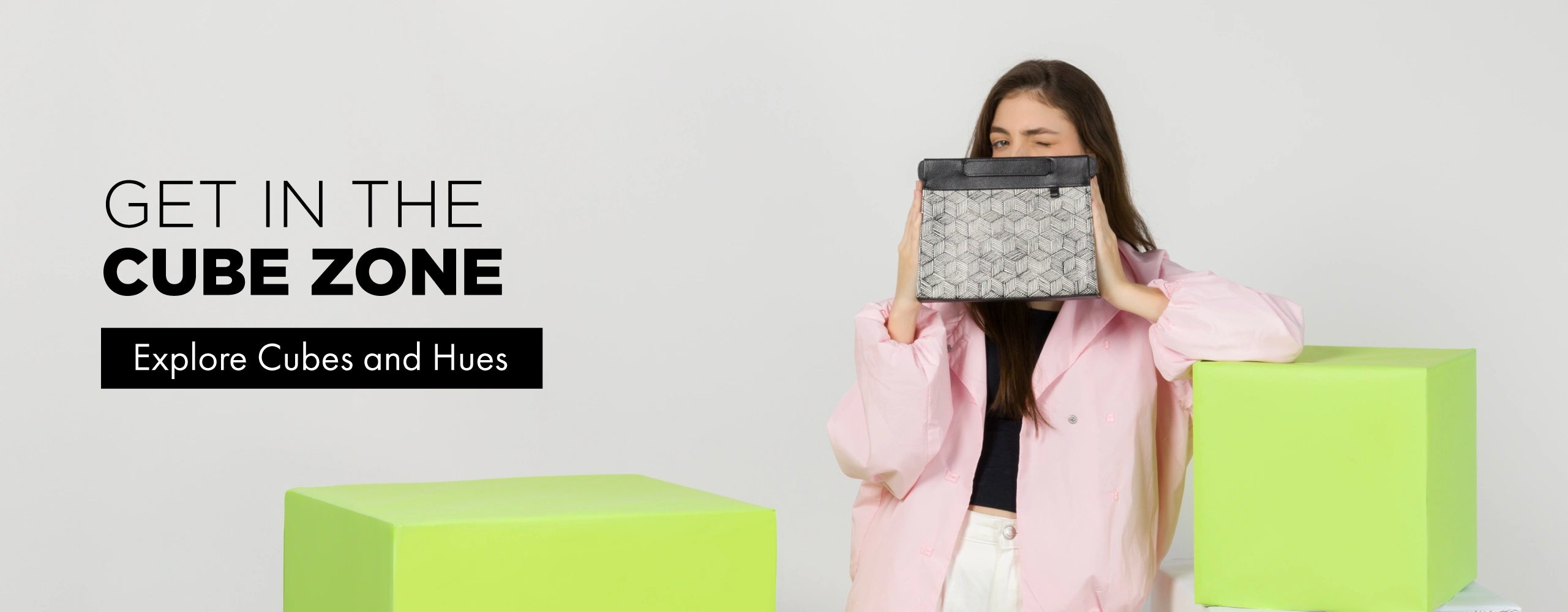 Woman winking while holding a black and white cube-print clutch bag up to her face, wearing a pink jacket over a black top and white pants, standing next to lime green cube-shaped boxes. Text reads "Get In The Cube Zone" with a button labeled "Explore Cubes and Hues."