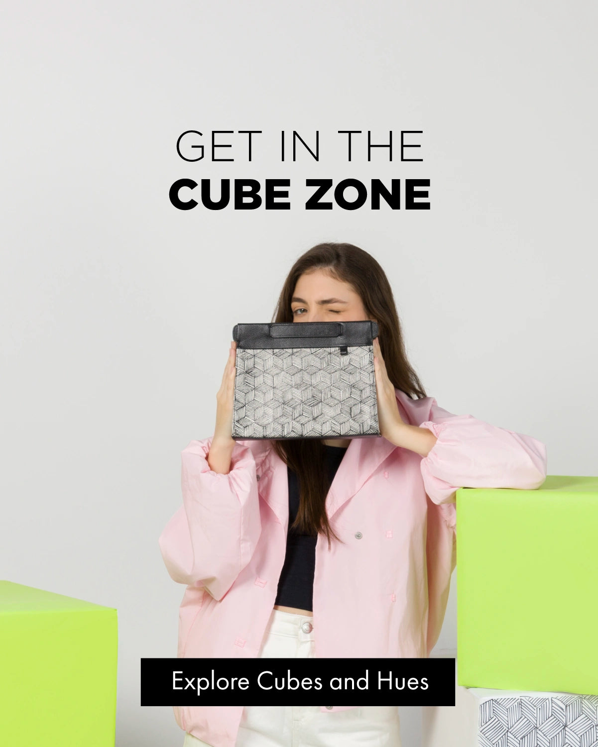 Woman winking while holding a black and white cube-print clutch bag up to her face, wearing a pink jacket over a black top and white pants, standing next to lime green cube-shaped boxes. Text reads "Get In The Cube Zone" with a button labeled "Explore Cubes and Hues."