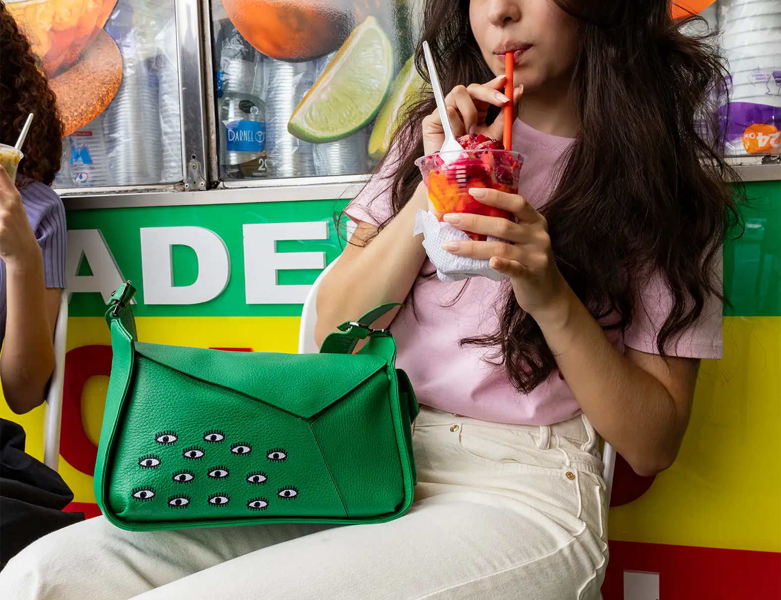 Young woman with long hair sipping a bright frozen drink through a straw in front of a colorful fruit stand. Large green shoulder bag on her lap. Relaxed vibe.
