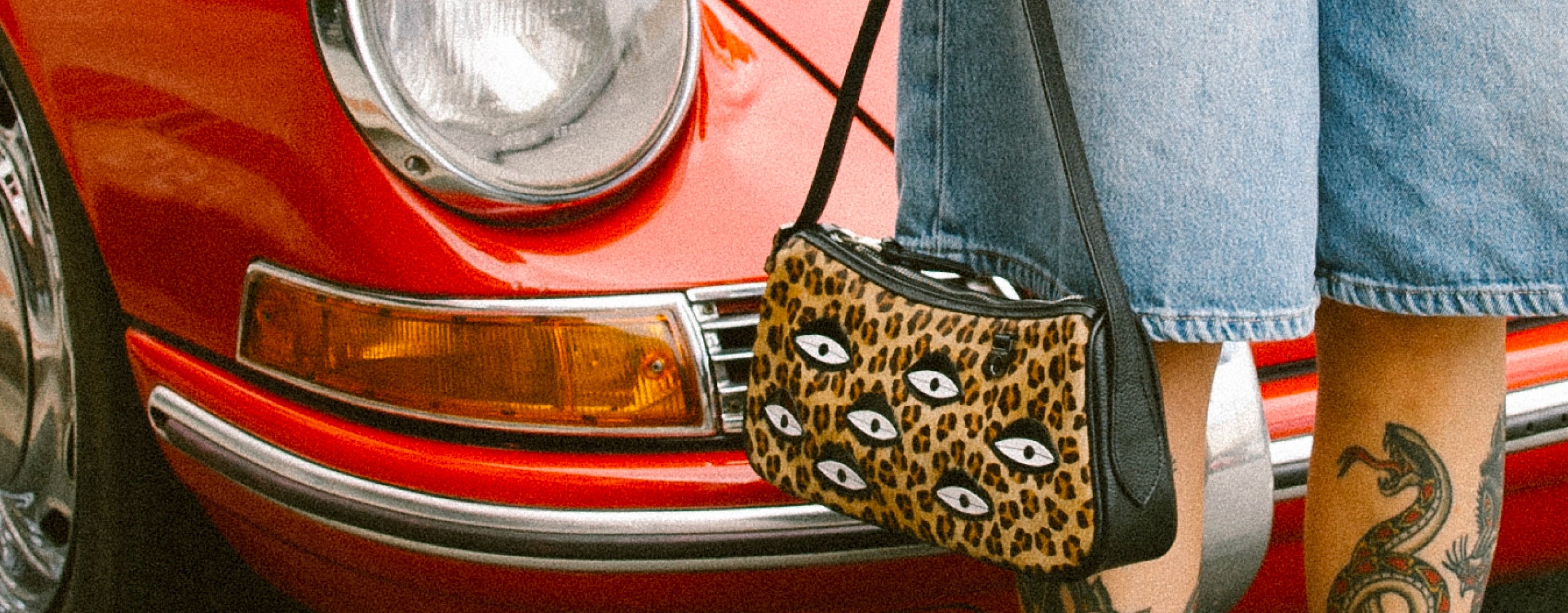 Tattoed woman in denim shorts and black boots holding a leopard-print eye-motif shoulder bag beside the headlight of a red vintage car.