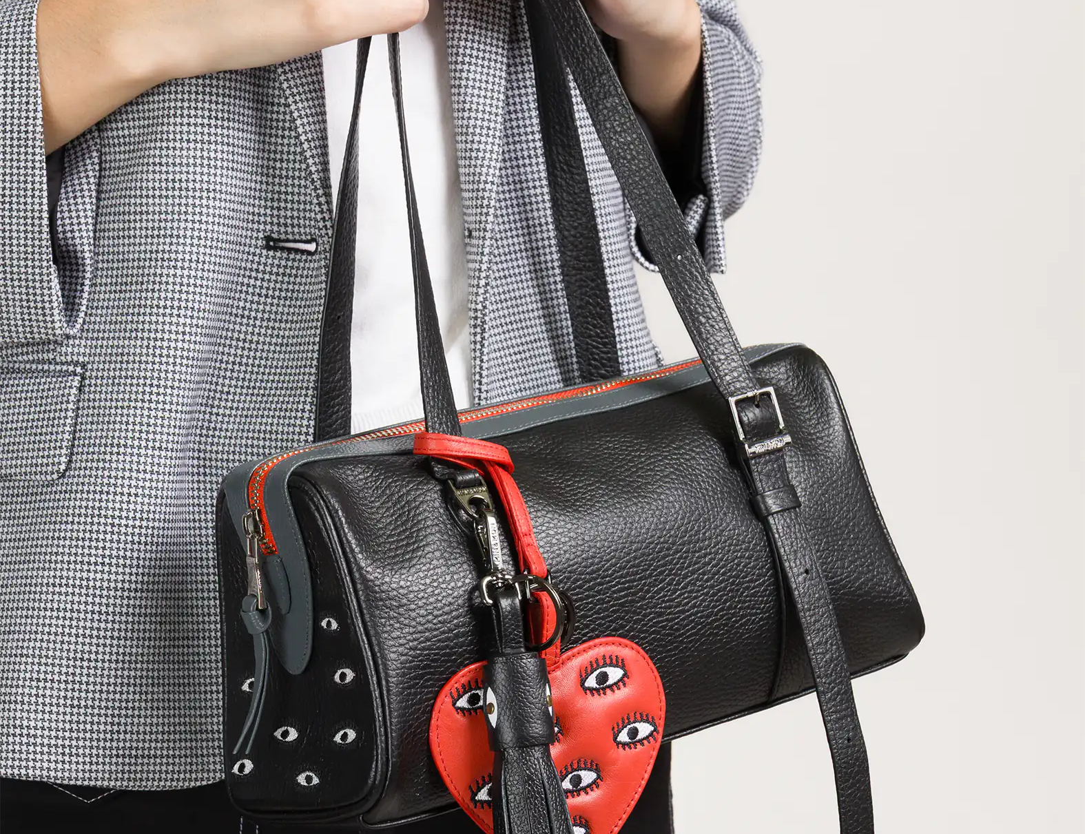 A person wearing a gray checkered blazer holds a black leather handbag adorned with a red heart-shaped charm featuring embroidered eyes and a black tassel.