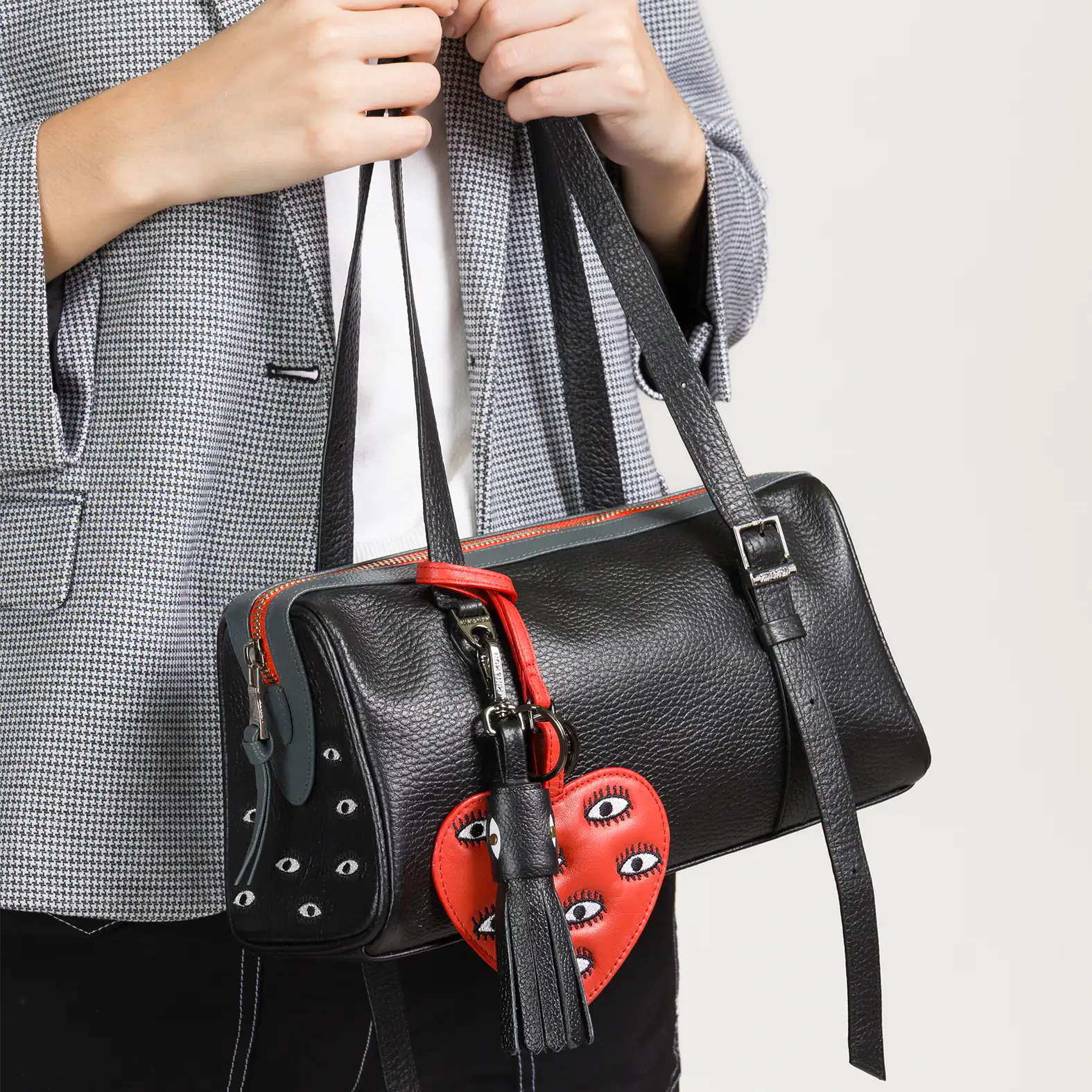 A person wearing a gray checkered blazer holds a black leather handbag adorned with a red heart-shaped charm featuring embroidered eyes and a black tassel.