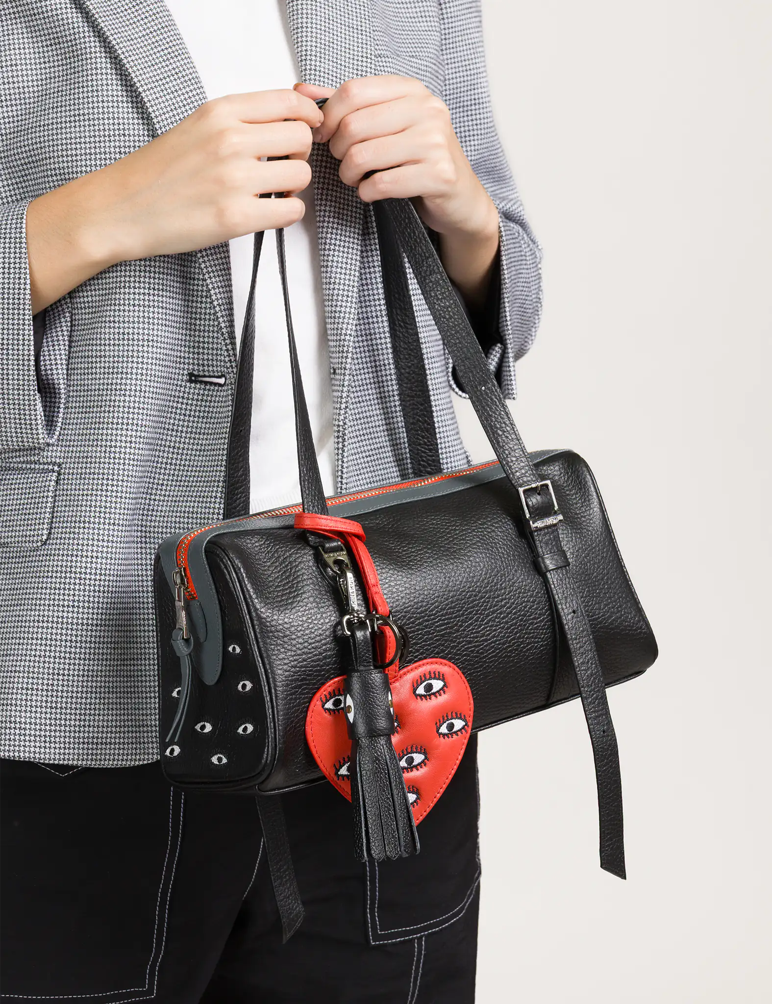 A person wearing a gray checkered blazer holds a black leather handbag adorned with a red heart-shaped charm featuring embroidered eyes and a black tassel.