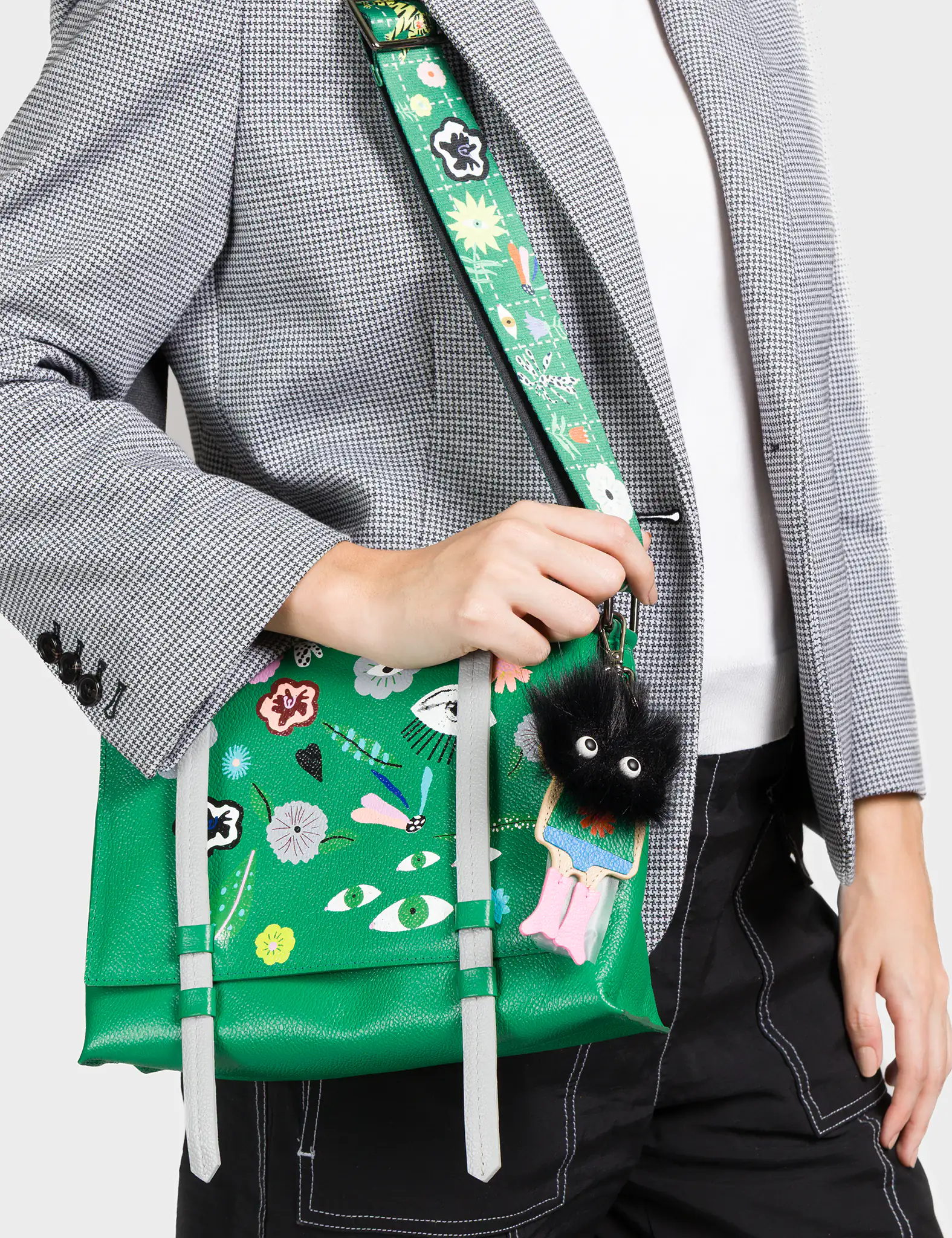 A woman wearing a black and white checkered blazer and black pants holds a vivid green handbag with whimsical embroidered details including eyes, flowers, and abstract motifs. The bag features two vertical light gray straps and a matching green shoulder strap with a playful illustrated pattern. Attached to the bag is a fuzzy black keychain charm with big round eyes and dangling pink and blue legs.