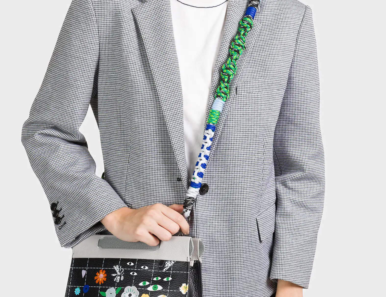 A smiling woman wearing a black and white checkered blazer over a white t-shirt carries a stylish crossbody bag. The bag features a black grid pattern adorned with colorful floral designs and whimsical eye motifs. It has a light gray flap and a bold, multicolored braided strap in green, blue, and white tones.