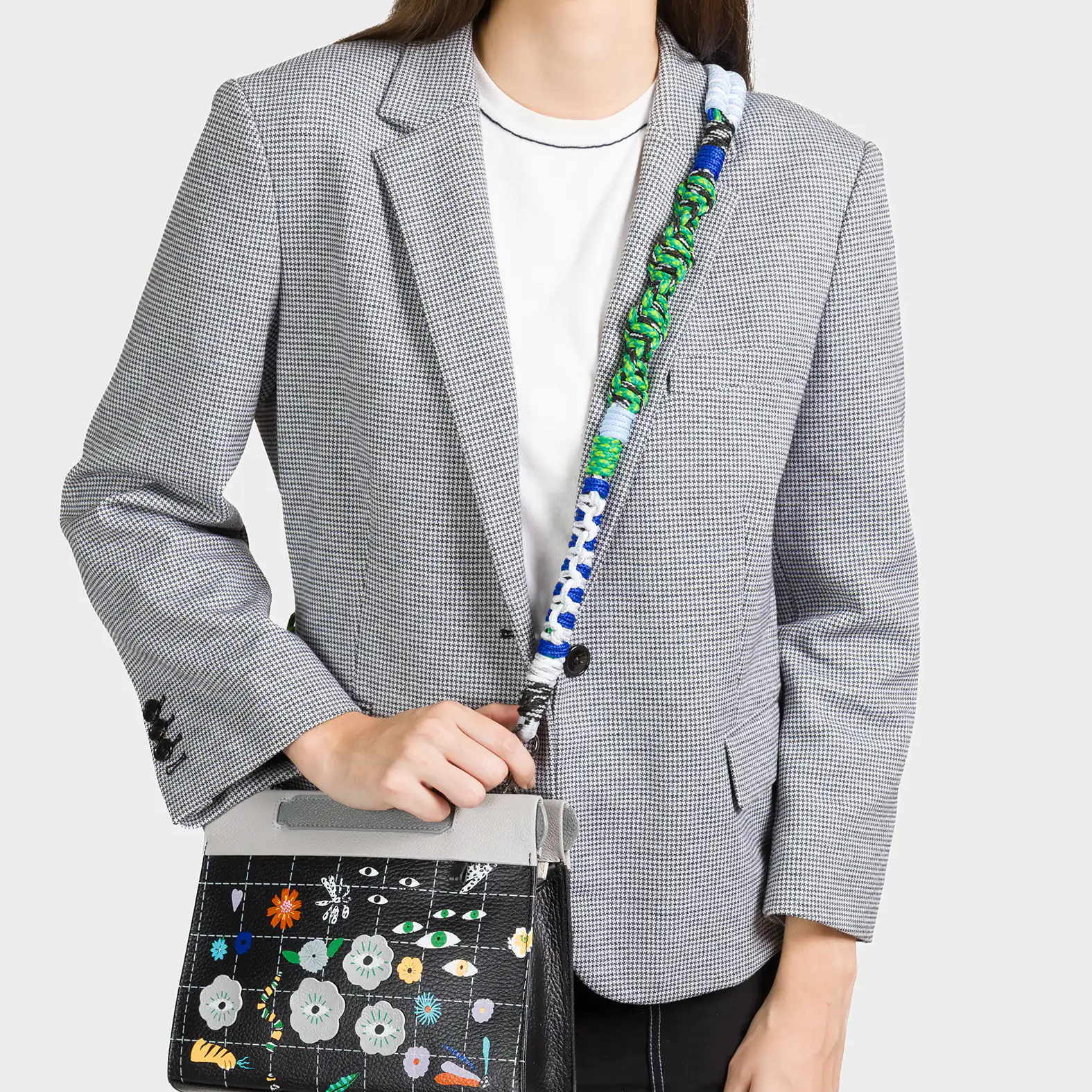 A smiling woman wearing a black and white checkered blazer over a white t-shirt carries a stylish crossbody bag. The bag features a black grid pattern adorned with colorful floral designs and whimsical eye motifs. It has a light gray flap and a bold, multicolored braided strap in green, blue, and white tones.