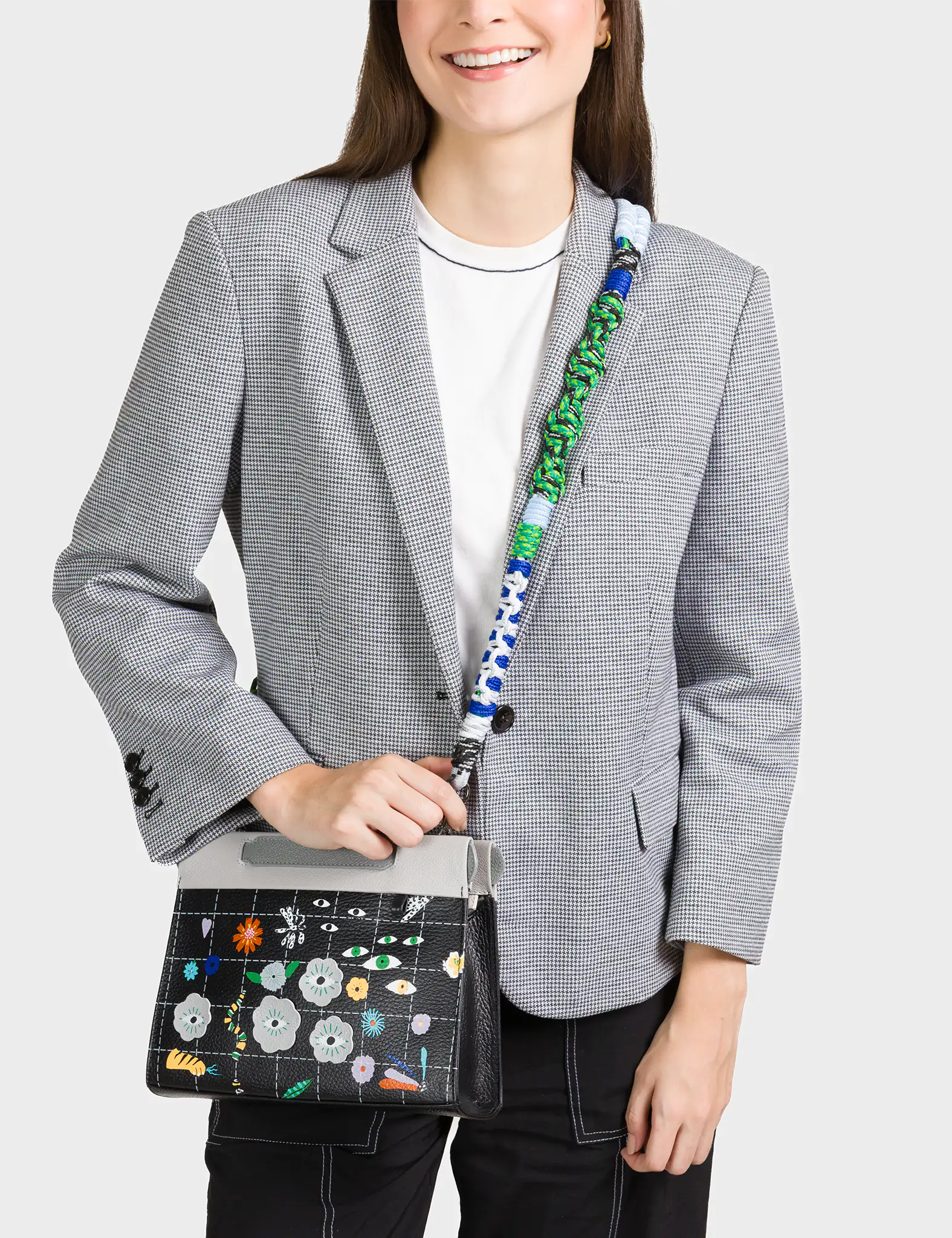 A smiling woman wearing a black and white checkered blazer over a white t-shirt carries a stylish crossbody bag. The bag features a black grid pattern adorned with colorful floral designs and whimsical eye motifs. It has a light gray flap and a bold, multicolored braided strap in green, blue, and white tones.