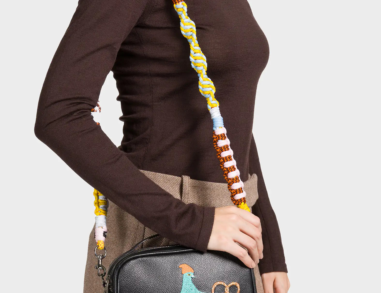A woman wearing a brown turtleneck top and a beige mini skirt carries a black crossbody bag. The bag features a colorful embroidered design of a blue pigeon wearing a beanie, holding a pretzel, and standing on top of a yellow taxi. It has a thick, multicolored woven shoulder strap in blue, yellow, brown, and white tones.