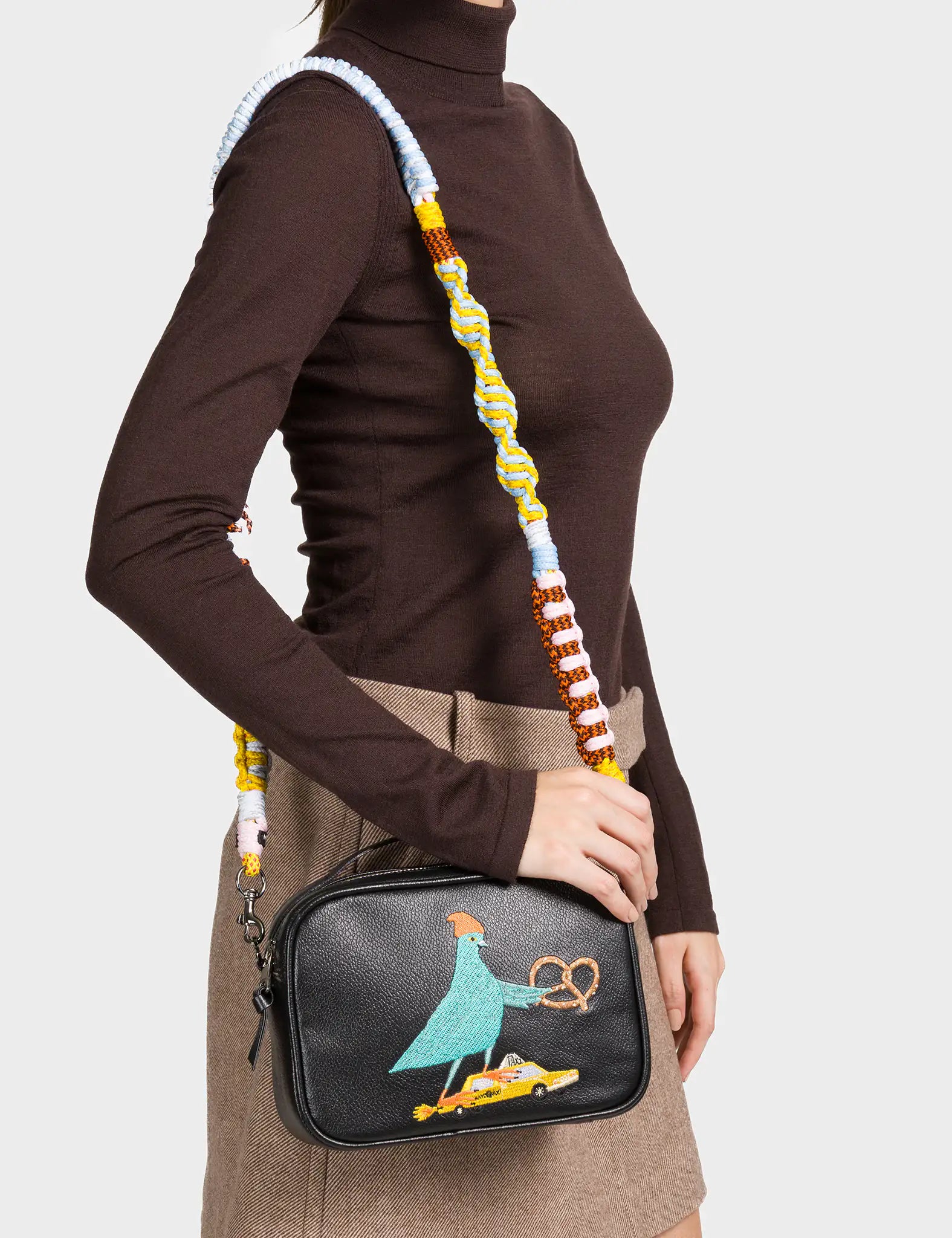 A woman wearing a brown turtleneck top and a beige mini skirt carries a black crossbody bag. The bag features a colorful embroidered design of a blue pigeon wearing a beanie, holding a pretzel, and standing on top of a yellow taxi. It has a thick, multicolored woven shoulder strap in blue, yellow, brown, and white tones.