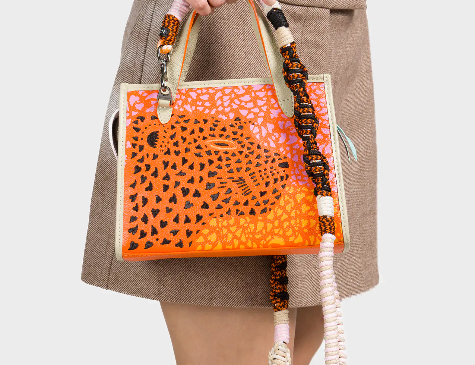 A woman wearing a brown long-sleeve top and a beige mini skirt holds a small structured handbag in her hand. The bag features an orange and pink abstract animal print design with a large spotted motif at the center. It has cream-colored edges and a chunky, multi-colored woven shoulder strap in shades of black, orange, pink, and white.