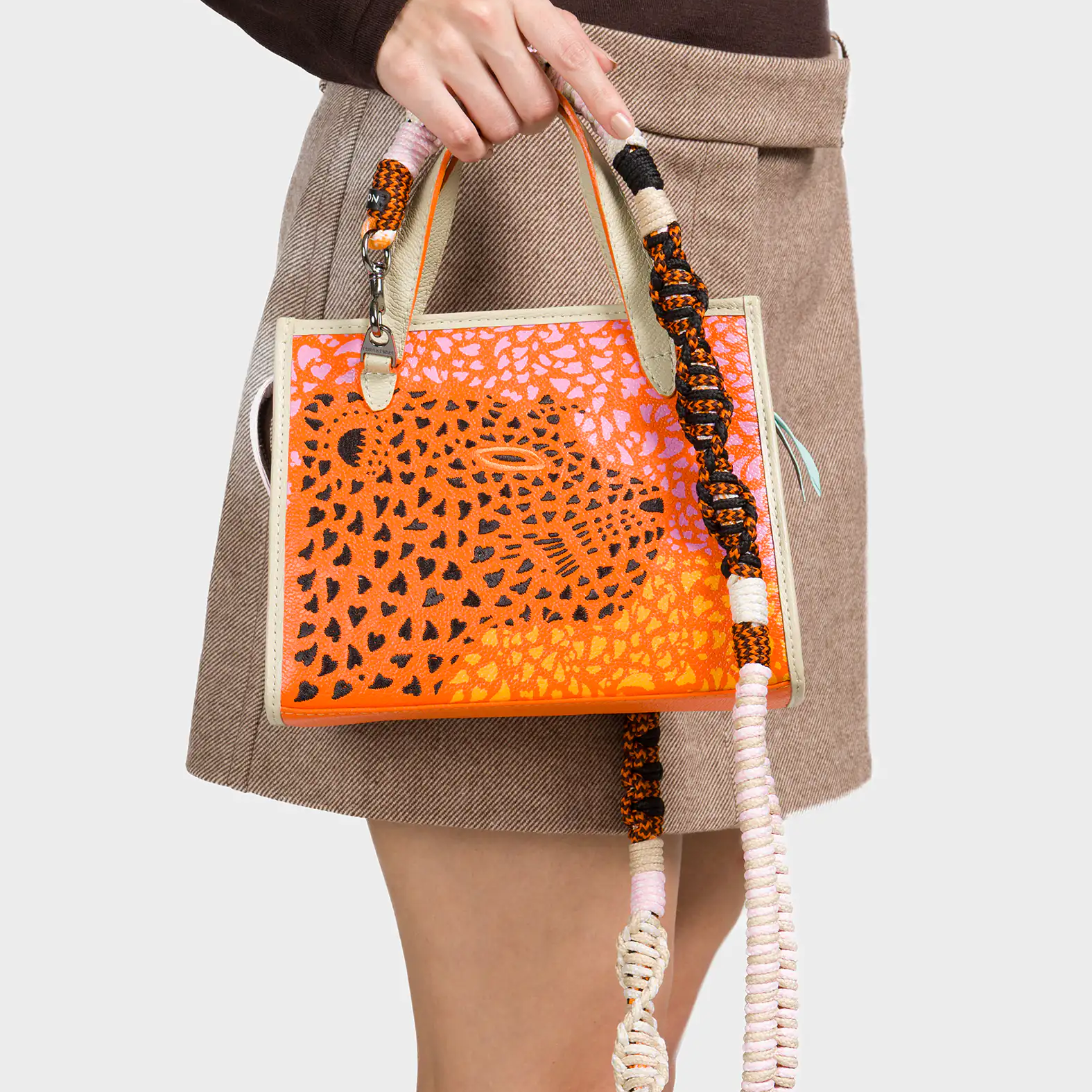 A woman wearing a brown long-sleeve top and a beige mini skirt holds a small structured handbag in her hand. The bag features an orange and pink abstract animal print design with a large spotted motif at the center. It has cream-colored edges and a chunky, multi-colored woven shoulder strap in shades of black, orange, pink, and white.