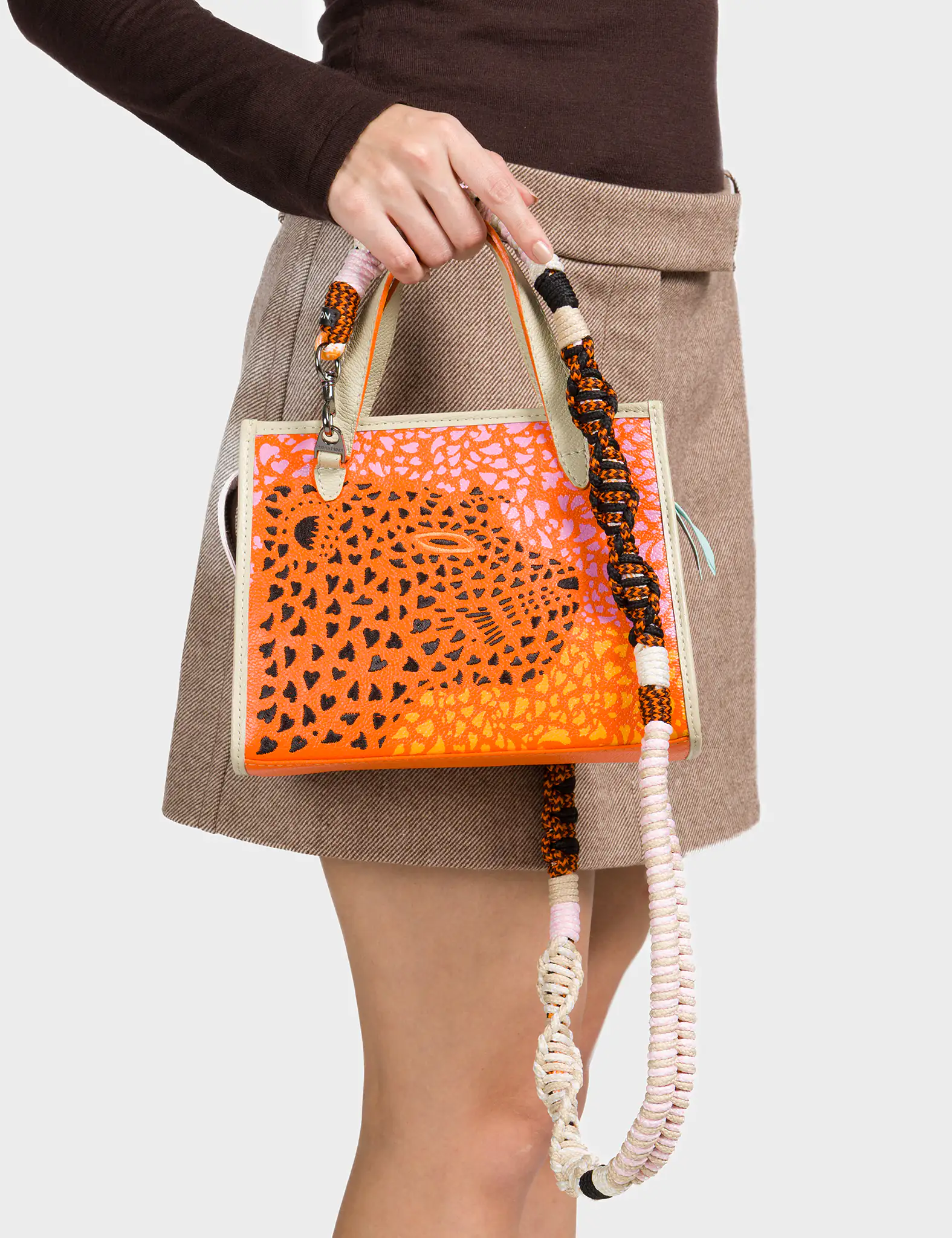 A woman wearing a brown long-sleeve top and a beige mini skirt holds a small structured handbag in her hand. The bag features an orange and pink abstract animal print design with a large spotted motif at the center. It has cream-colored edges and a chunky, multi-colored woven shoulder strap in shades of black, orange, pink, and white.