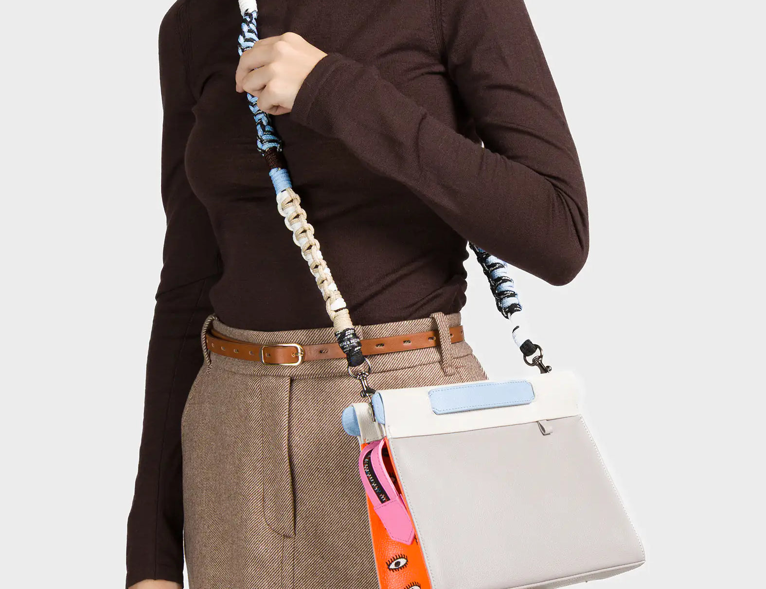 A woman dressed in a brown turtleneck and beige mini skirt with a brown belt carries a structured cream-colored handbag with a light blue flap detail. The bag features a whimsical side accent in orange and pink with illustrated eyes and includes a multicolored, twisted woven strap in shades of blue, black, and cream.