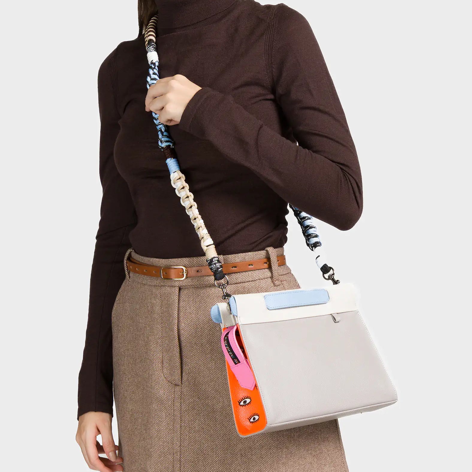 A woman dressed in a brown turtleneck and beige mini skirt with a brown belt carries a structured cream-colored handbag with a light blue flap detail. The bag features a whimsical side accent in orange and pink with illustrated eyes and includes a multicolored, twisted woven strap in shades of blue, black, and cream.