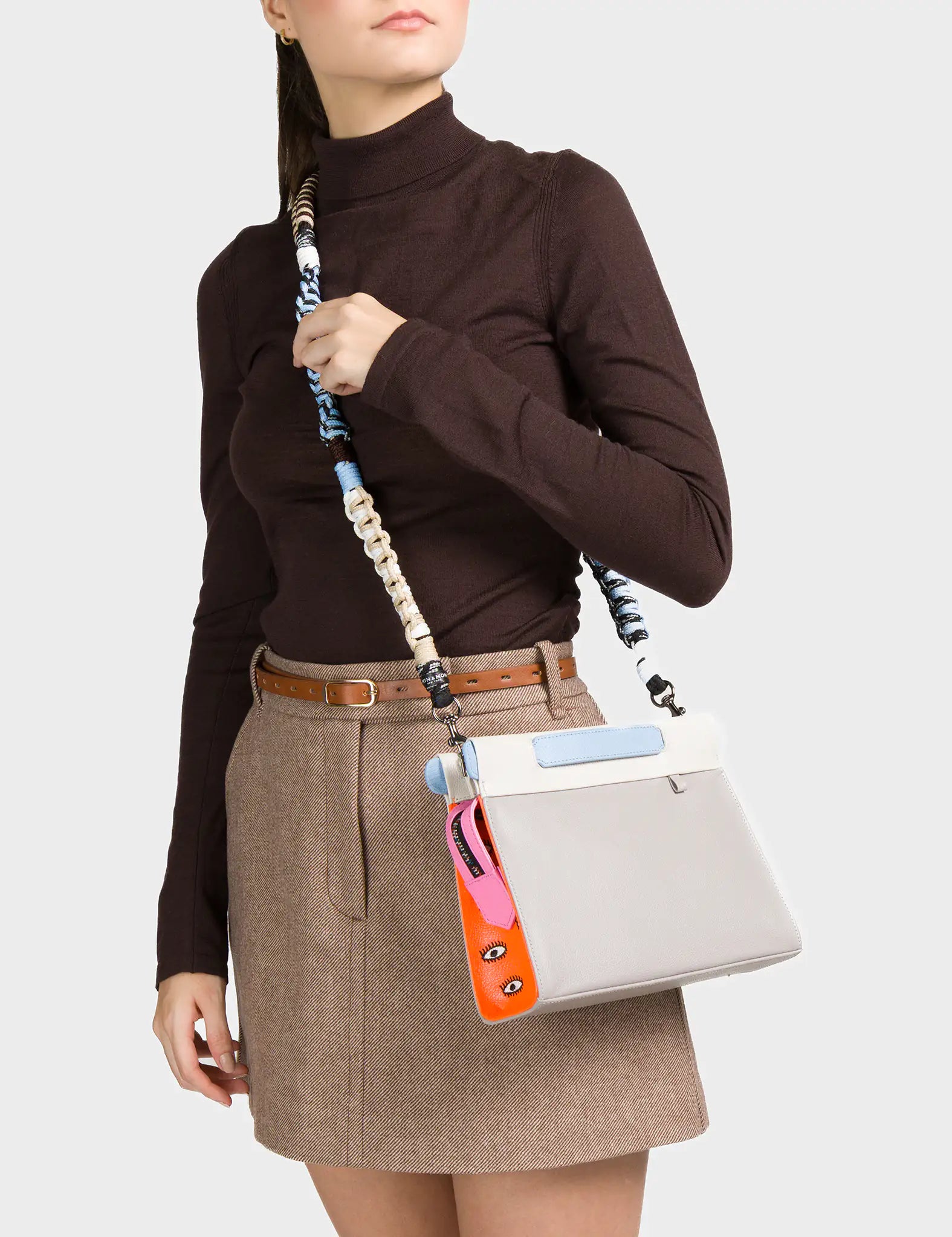 A woman dressed in a brown turtleneck and beige mini skirt with a brown belt carries a structured cream-colored handbag with a light blue flap detail. The bag features a whimsical side accent in orange and pink with illustrated eyes and includes a multicolored, twisted woven strap in shades of blue, black, and cream.