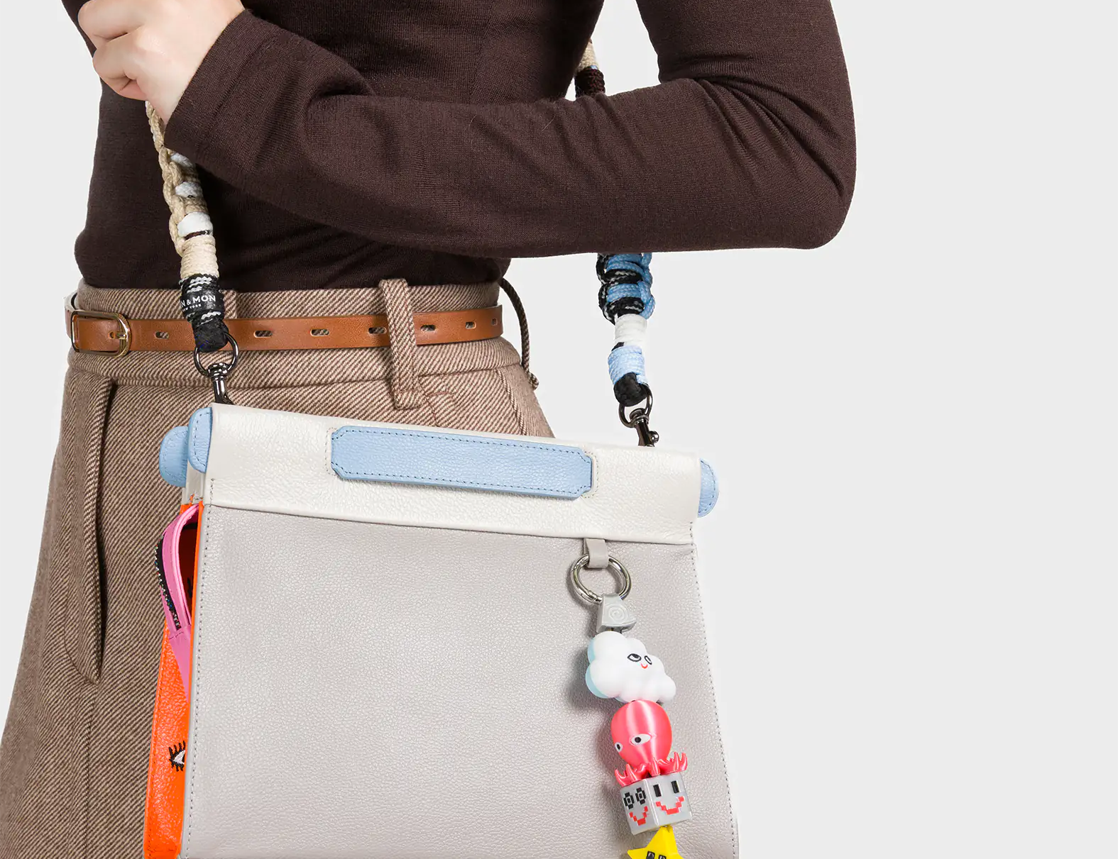 A woman dressed in a brown long-sleeve top and beige skirt carries a light gray structured handbag with baby blue and white trim. The bag features a colorful charm dangling from the strap, including a smiling cloud, a red octopus, a dice block, and a yellow star. A pink and orange accent tag with illustrated eyes peeks out from the side, adding a quirky and playful touch.