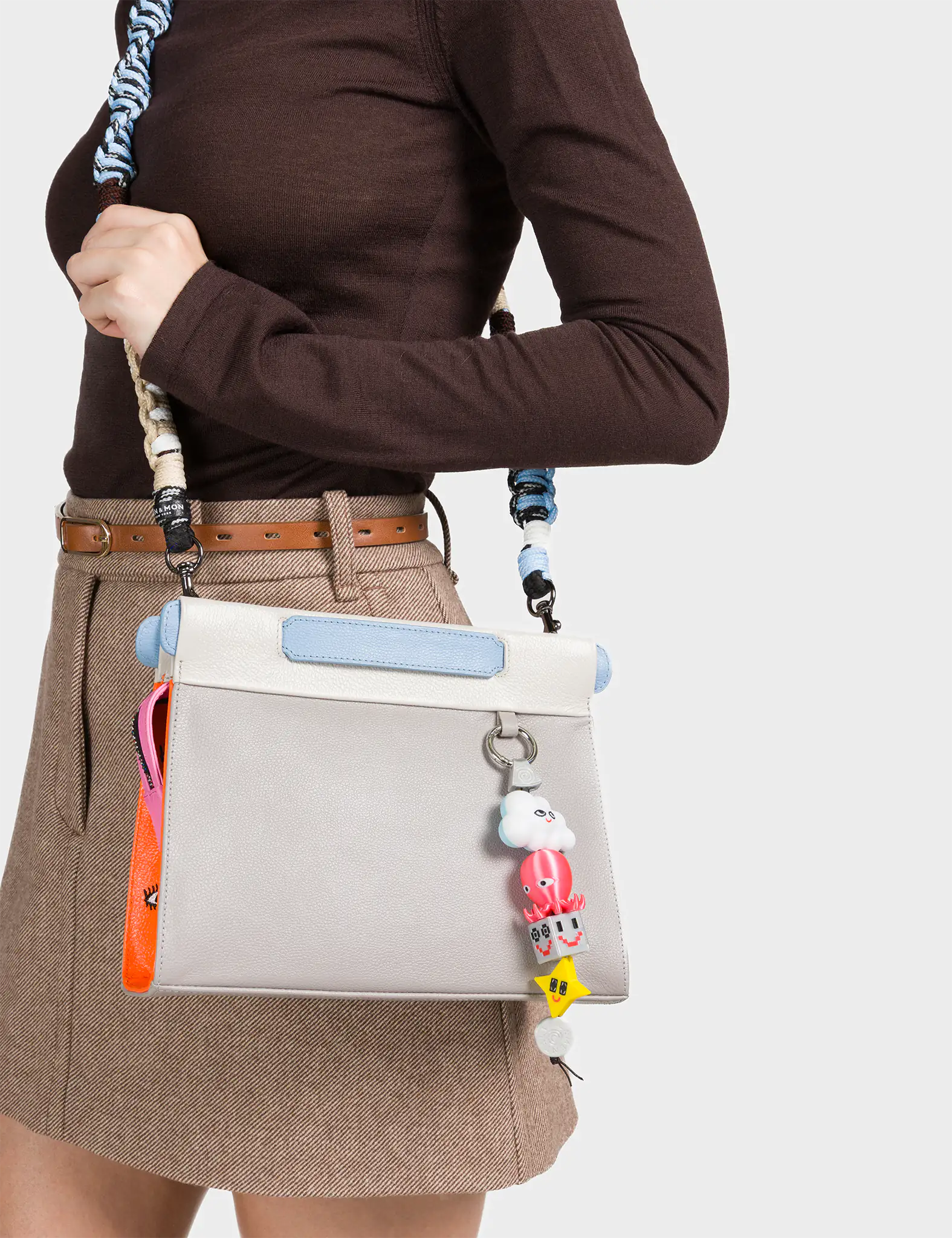 A woman dressed in a brown long-sleeve top and beige skirt carries a light gray structured handbag with baby blue and white trim. The bag features a colorful charm dangling from the strap, including a smiling cloud, a red octopus, a dice block, and a yellow star. A pink and orange accent tag with illustrated eyes peeks out from the side, adding a quirky and playful touch.