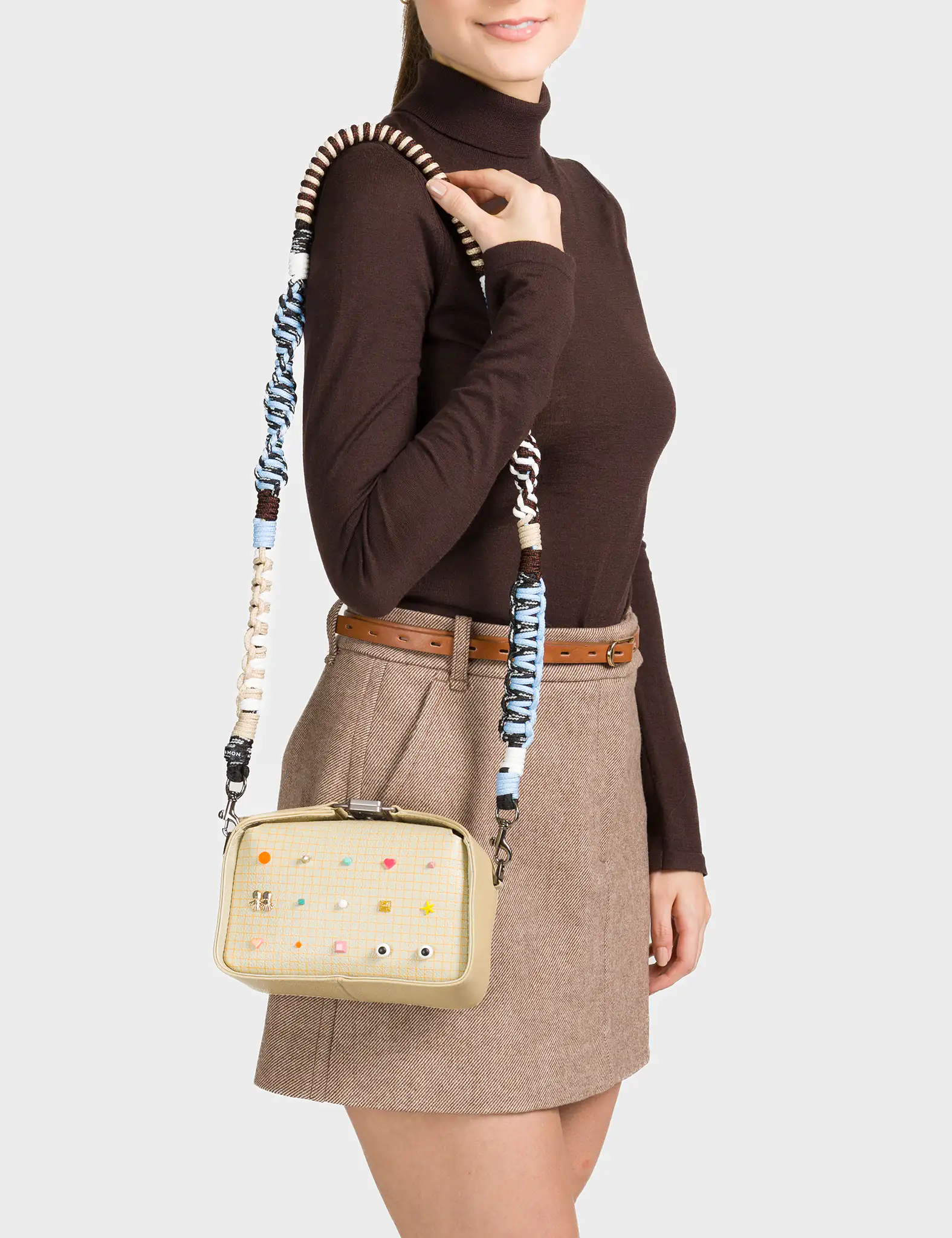 A woman wearing a brown turtleneck and beige mini skirt with a brown belt carries a small rectangular crossbody bag in a light beige tone. The front of the bag features a playful grid pattern decorated with colorful geometric icons and small illustrated eyes. The wide strap is intricately woven in shades of blue, cream, and brown.