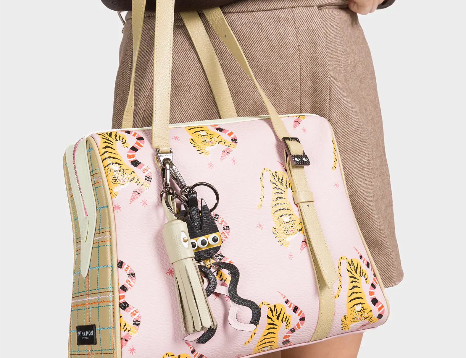 A woman wearing a brown long-sleeve top and beige skirt carries a pastel pink handbag decorated with playful yellow tigers and pink squiggles. The structured bag features beige plaid side panels and matching beige handles. A cluster of quirky keychains, including a white tassel and a small creature with googly eyes, hangs from one handle, adding a fun and whimsical accent.