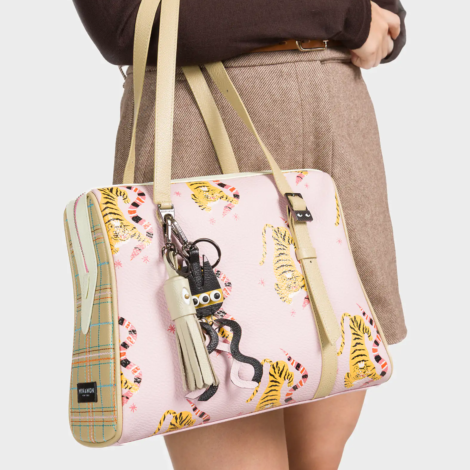 A woman wearing a brown long-sleeve top and beige skirt carries a pastel pink handbag decorated with playful yellow tigers and pink squiggles. The structured bag features beige plaid side panels and matching beige handles. A cluster of quirky keychains, including a white tassel and a small creature with googly eyes, hangs from one handle, adding a fun and whimsical accent.