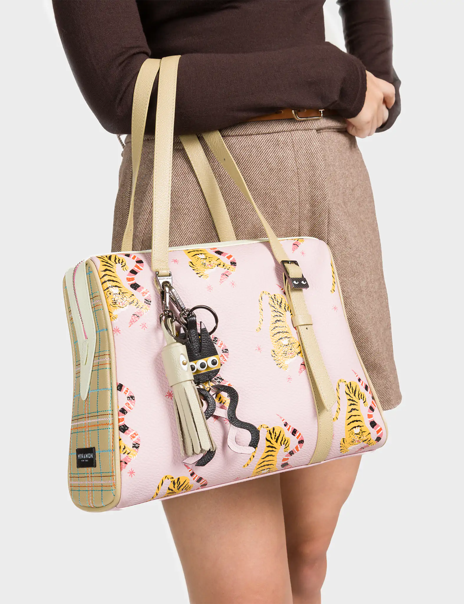 A woman wearing a brown long-sleeve top and beige skirt carries a pastel pink handbag decorated with playful yellow tigers and pink squiggles. The structured bag features beige plaid side panels and matching beige handles. A cluster of quirky keychains, including a white tassel and a small creature with googly eyes, hangs from one handle, adding a fun and whimsical accent.