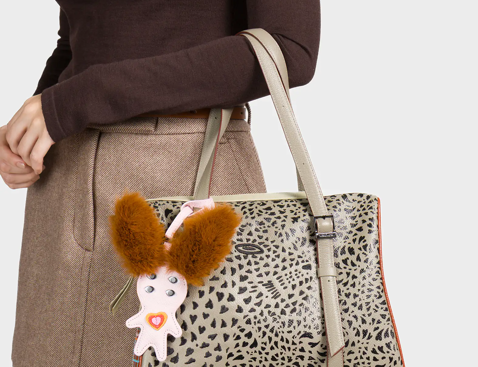 A woman dressed in a brown long-sleeve top and beige mini skirt holds a structured taupe handbag with an abstract black animal-print pattern. The bag features double handles and a vertical strap detail. A quirky charm shaped like a creature with large fluffy brown ears and a pink body is attached to one handle, adding a playful touch.