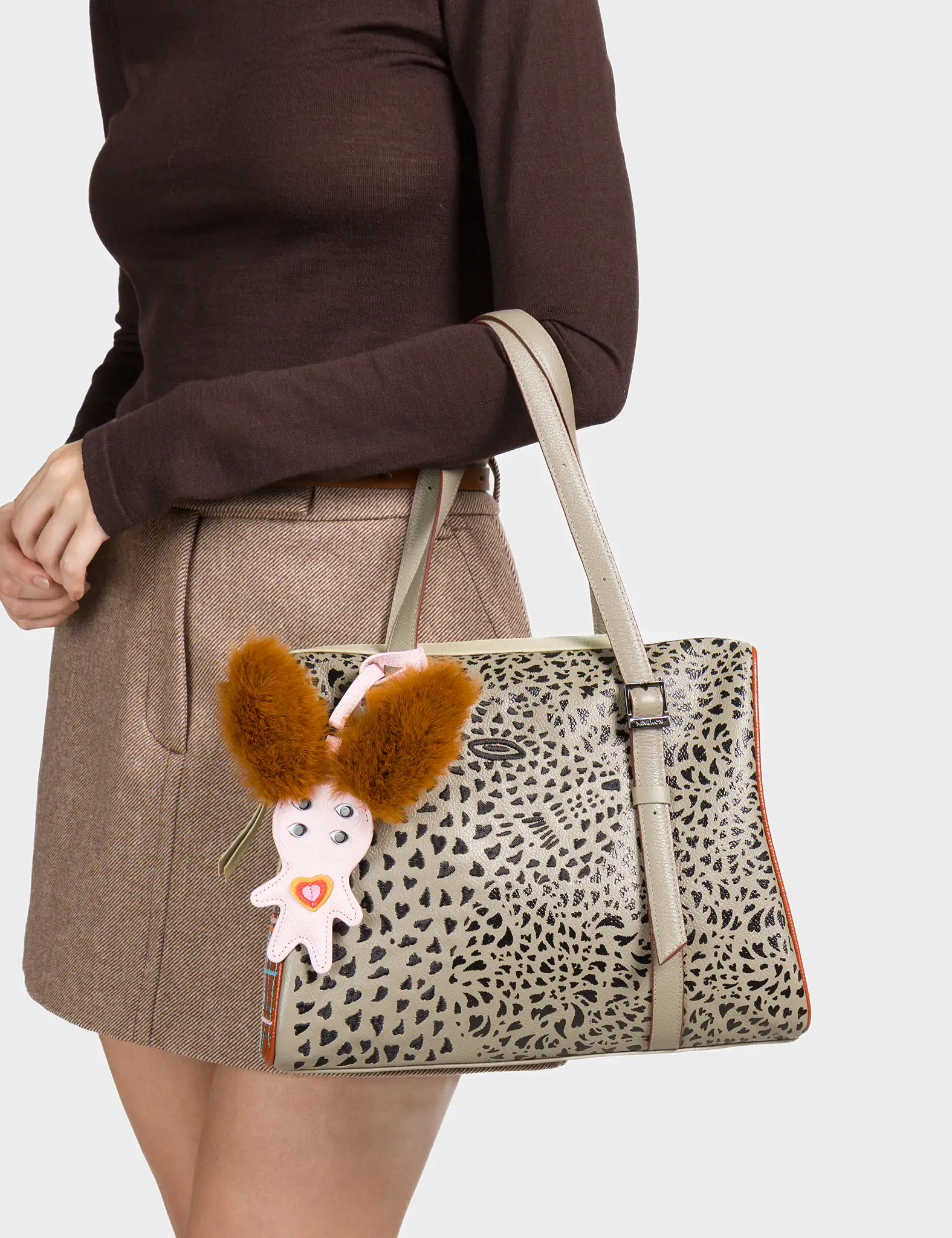 A woman dressed in a brown long-sleeve top and beige mini skirt holds a structured taupe handbag with an abstract black animal-print pattern. The bag features double handles and a vertical strap detail. A quirky charm shaped like a creature with large fluffy brown ears and a pink body is attached to one handle, adding a playful touch.