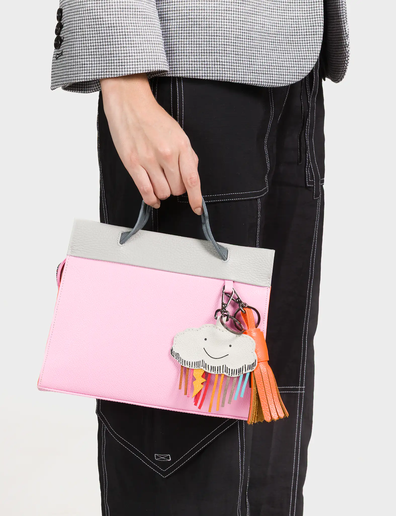A person in a checkered blazer and black pants with white stitching holds a pastel pink and light gray structured handbag. Attached to the handle is a cheerful cloud-shaped charm with colorful rainbow fringe and a lightning bolt, accompanied by a textured orange tassel keychain, adding a fun and whimsical touch to the look.
