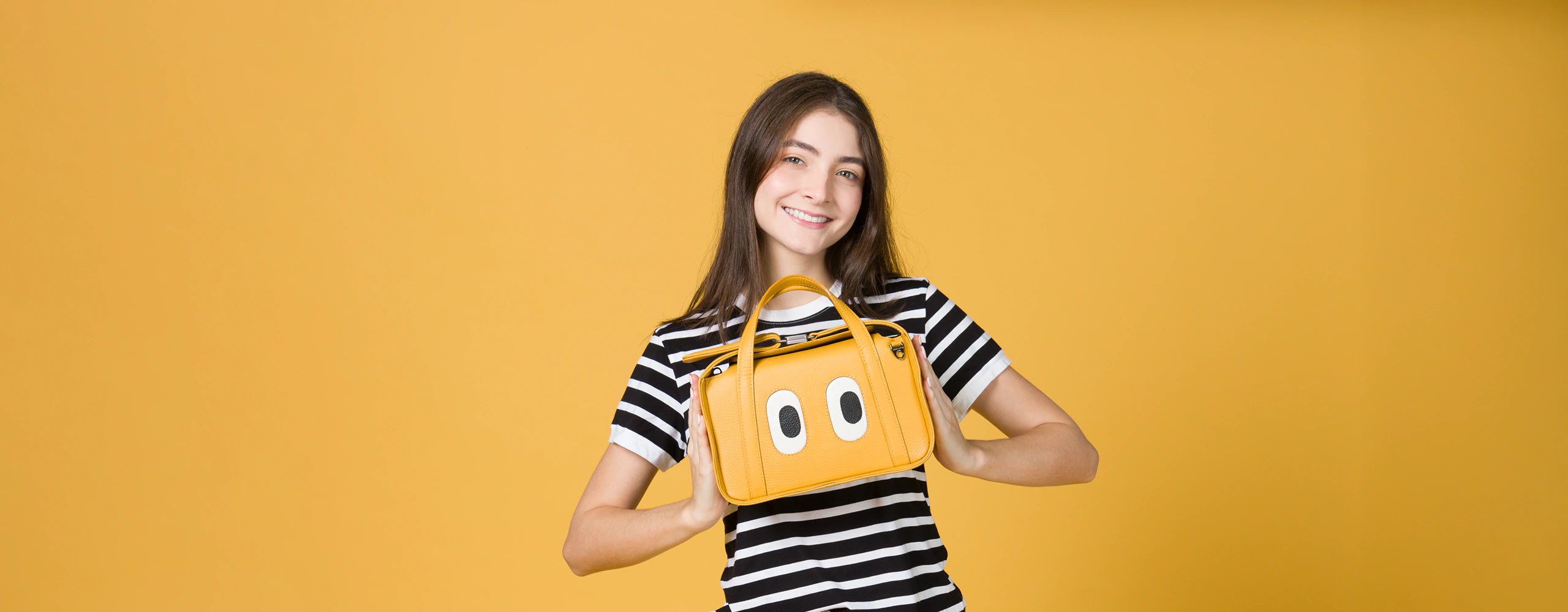 Young woman smiling while holding a yellow Min & Mon handbag with eye appliqués, against a bright yellow background.