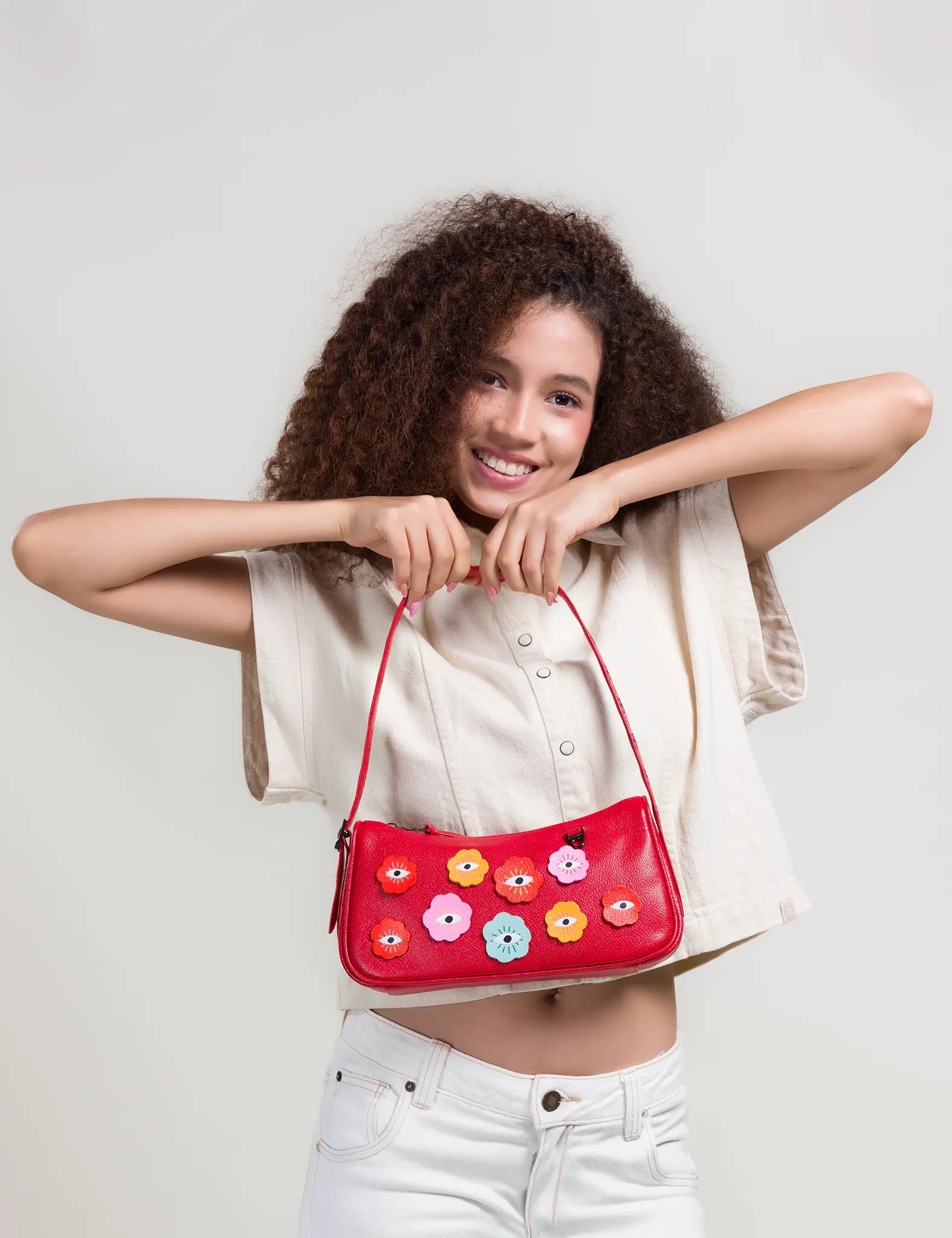 Smiling woman with curly hair holds a red shoulder bag featuring colorful eye motifs, wearing a light top and white pants against a neutral background.
