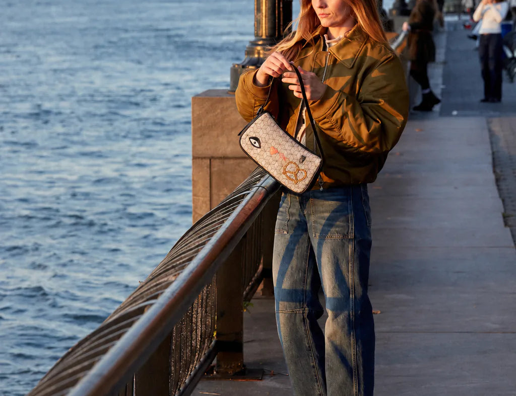 Woman in an olive jacket stands on a golden-hour waterfront promenade, adjusting a light patterned shoulder bag with eye, heart, and pretzel motifs.