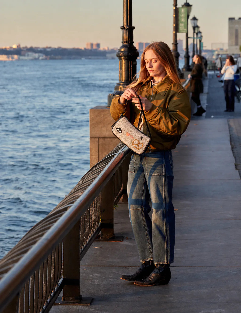 Woman in an olive jacket stands on a golden-hour waterfront promenade, adjusting a light patterned shoulder bag with eye, heart, and pretzel motifs.