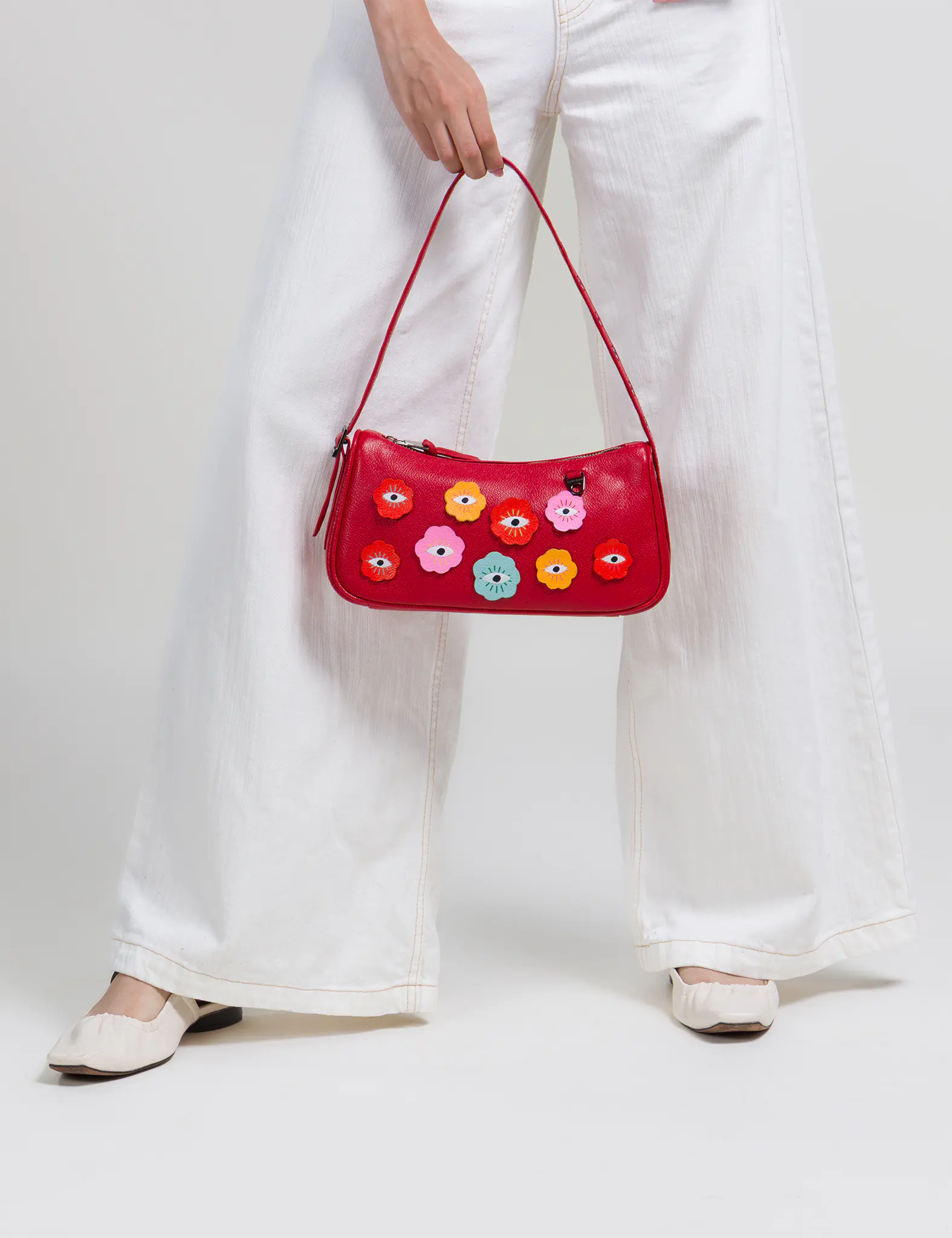 Close-up of a red shoulder bag with colorful eye designs, held by a person in white wide-leg pants and cream flats against a neutral studio backdrop.
