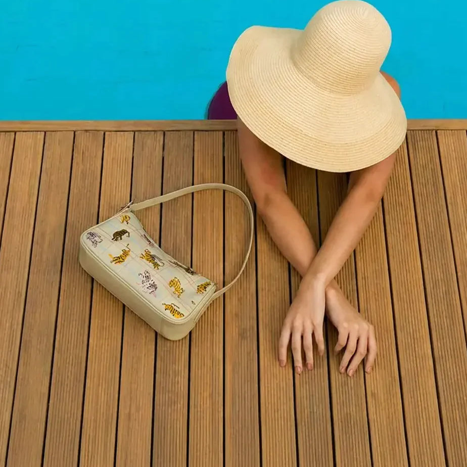 Top view of a woman in a wide-brim hat leaning over a pool edge, with a cream shoulder bag featuring tiger prints resting on a wooden deck beside her.