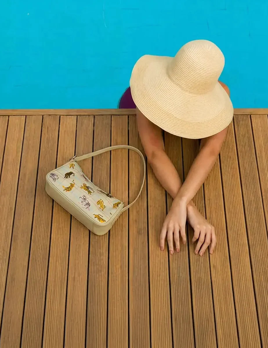 Top view of a woman in a wide-brim hat leaning over a pool edge, with a cream shoulder bag featuring tiger prints resting on a wooden deck beside her.